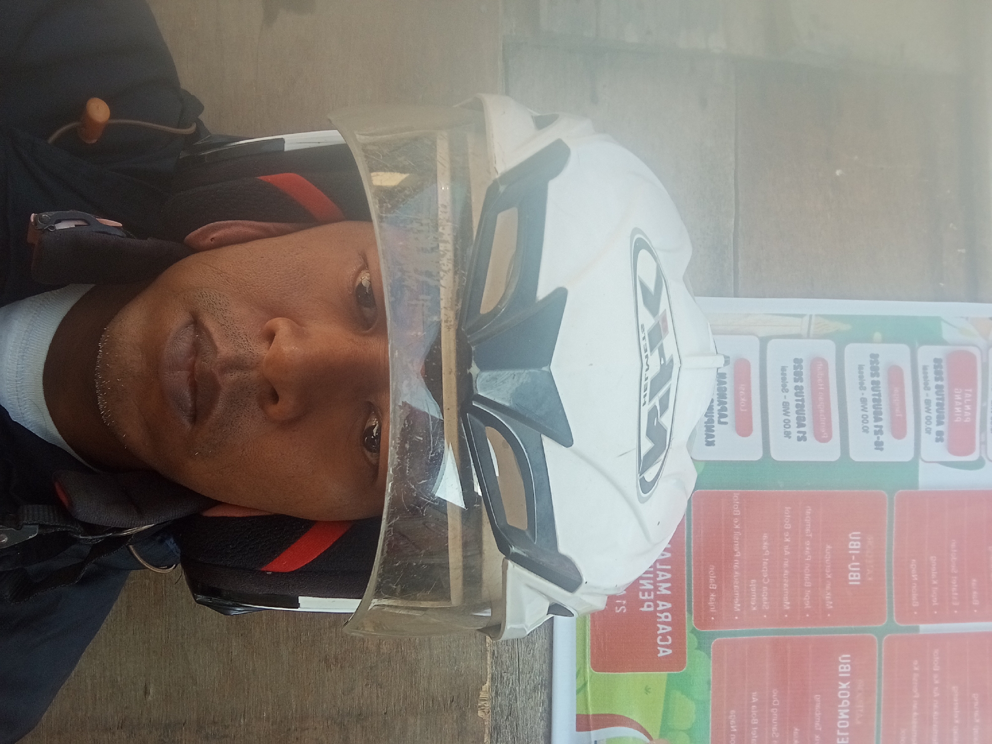 A person wearing a white and black motorcycle helmet with a clear visor is looking directly at the camera. The helmet has an "X" logo on it. The person's face is visible from the eyes down to the chin. They are wearing a dark jacket over a white shirt. In the background, there is a poster with text in Indonesian. The poster is positioned behind the person's head. The environment appears to be outdoors, with a wooden surface visible in the lower right of the image, suggesting it might be a public space or a street in Pagar Alam, Indonesia.