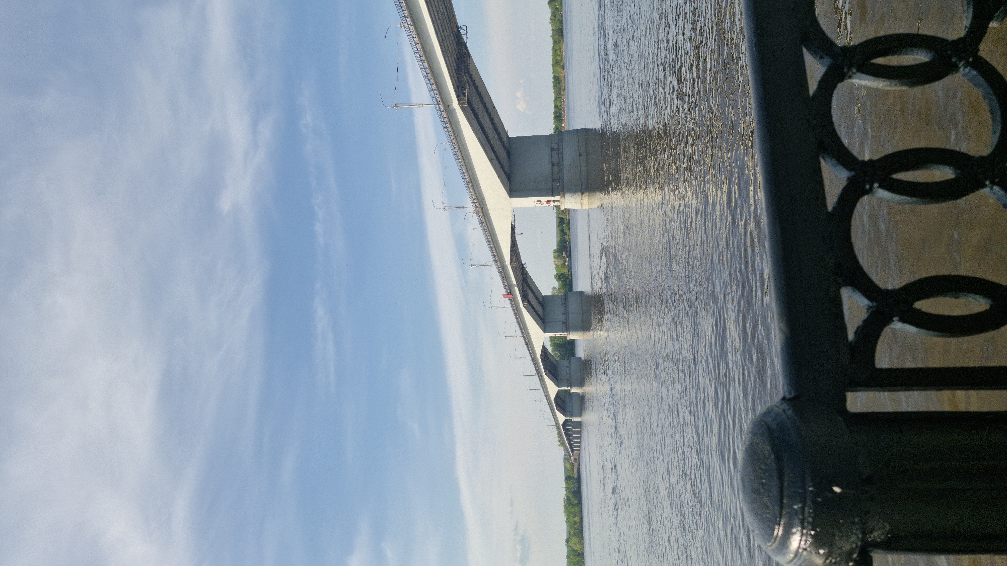An eye-level shot of the Kama River Bridge in Perm, Russia, is taken from a slightly elevated position, possibly a promenade or embankment. The bridge extends horizontally across the water, connecting the riverbanks. The image is divided into three main areas: sky, water, and bridge. The sky, visible on the left, is partly cloudy with a blue hue. The water of the Kama River occupies the central portion, displaying gentle ripples. The bridge's structure is gray concrete. On the right, a dark metal fence with a pattern of interlocking circles adds a layer of depth and detail.