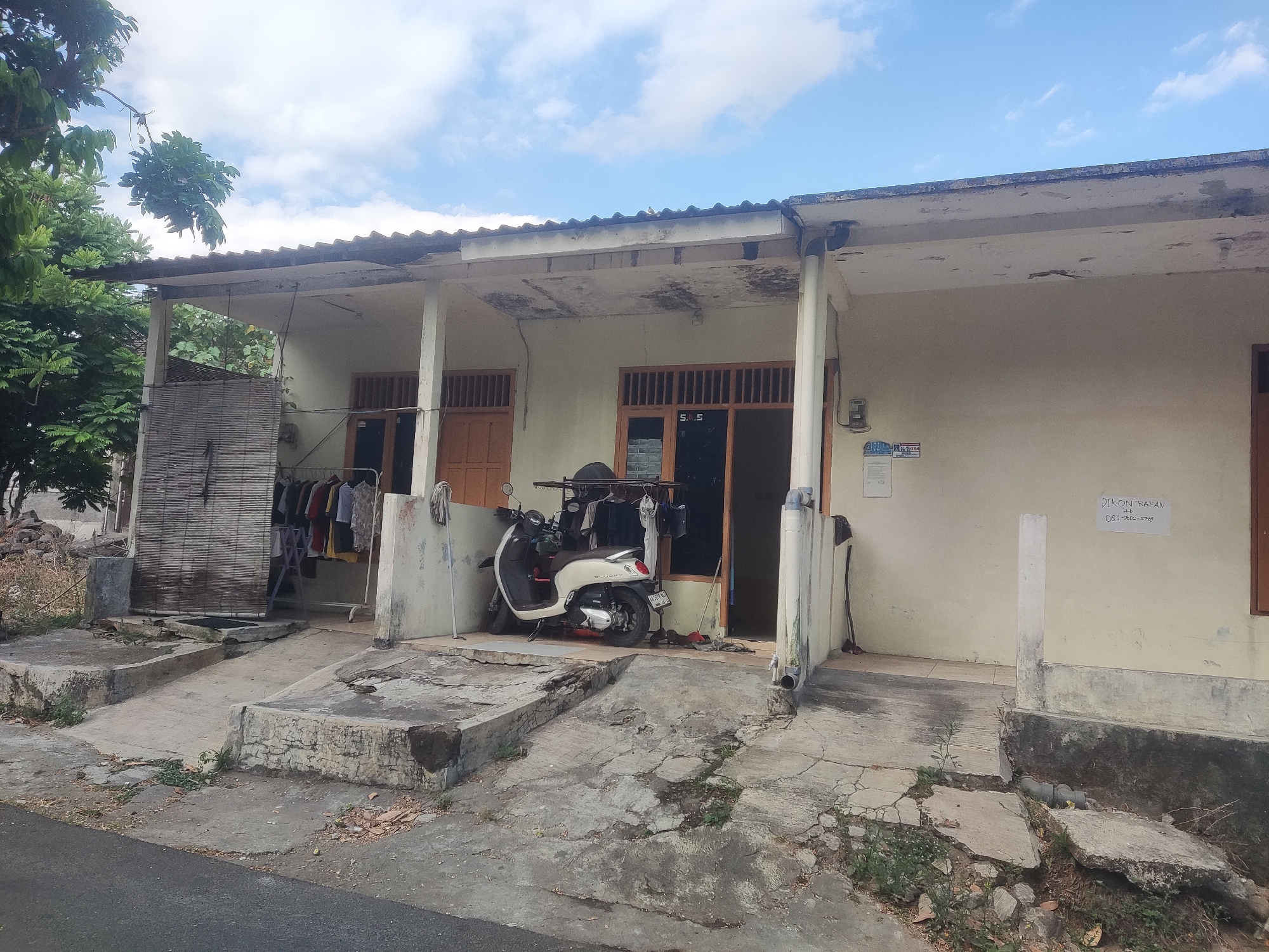The image depicts the exterior of a modest, single-story house with a covered porch. The house has a light-colored facade and a corrugated roof. In front of the house, there is a paved driveway with some cracks and vegetation growing through. A white scooter is parked on the driveway, and clothes are hanging on a rack to the left of the scooter. A bamboo blind is also visible to the left of the clothes rack. The environment appears to be a residential area, possibly in a tropical climate, with trees and a clear blue sky with some clouds. There are no people visible in the image. A sign on the wall to the right advertises a rental property with a phone number.