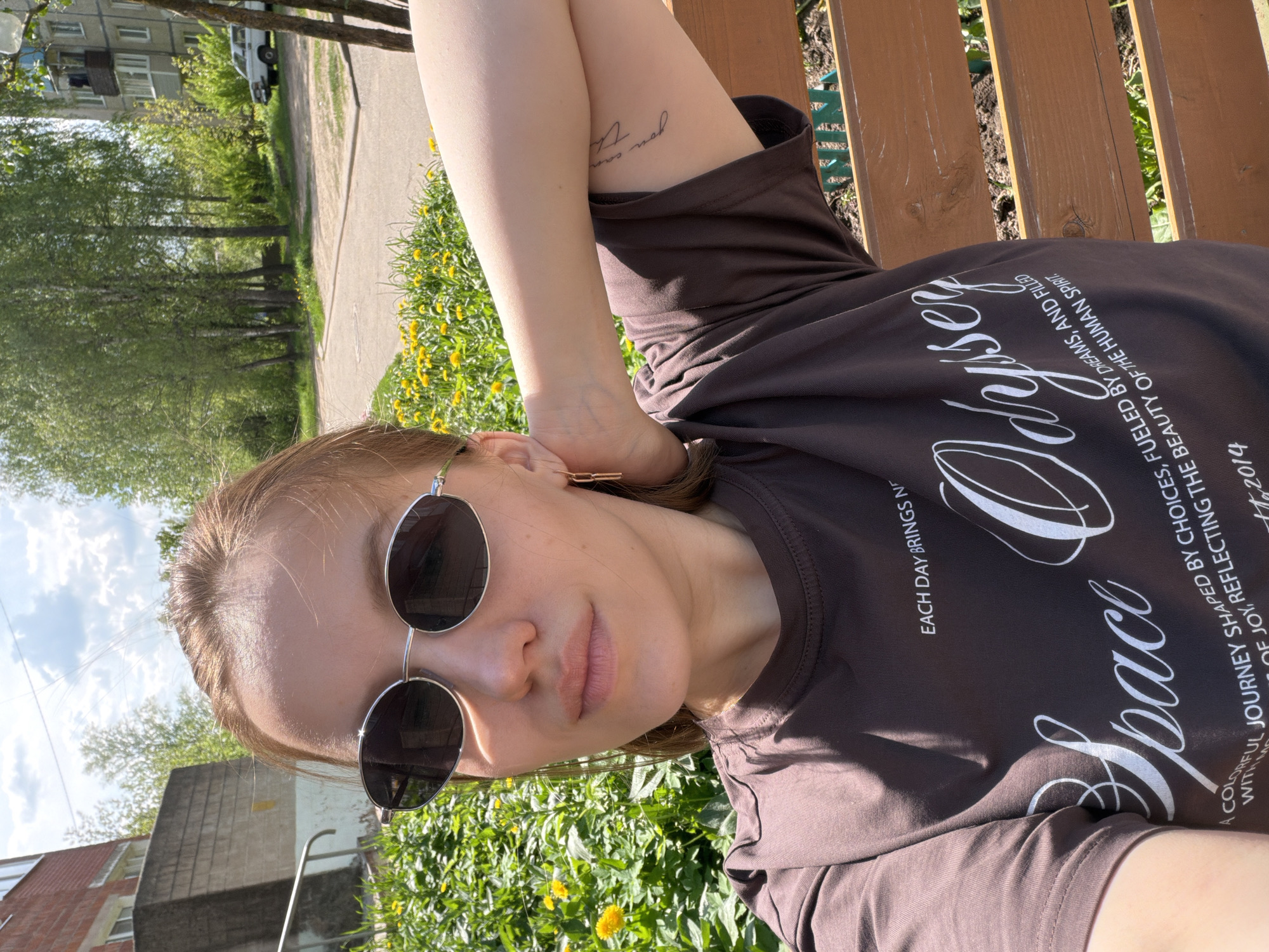 The image is a rotated portrait-style photograph of a young woman resting outdoors on a sunny day in Reshetnikovo, Russia. She is seen from the chest up, reclining, with her right arm bent and resting behind her head. She wears round, dark sunglasses and a dark brown t-shirt. Her light brown hair is pulled back, and a small, delicate, light-colored earring is visible in her right ear. On her inner right forearm, a small script tattoo is visible, which appears to read "you can" or a similar phrase.
Her t-shirt features white text, including "Space Odyssey" in a decorative font, and smaller text that reads "EACH DAY BRINGS NE", "A COLORFUL JOURNEY SHAPED BY CHOICES, FUELED BY DREAMS, AND FILLED WITH MO OF JOY, REFLECTING THE BEAUTY OF THE HUMAN SPIRIT.", and "112014".
The background suggests an urban or suburban green space. Bright sunlight illuminates the scene, and the sky above shows scattered clouds. Immediately behind her is a grassy area filled with yellow dandelions. Further back, a paved path or road runs alongside several tall, slender trees with lush green leaves. In the distant background, multi-story apartment buildings are visible, with a white vehicle parked nearby. The overall atmosphere is relaxed and peaceful.