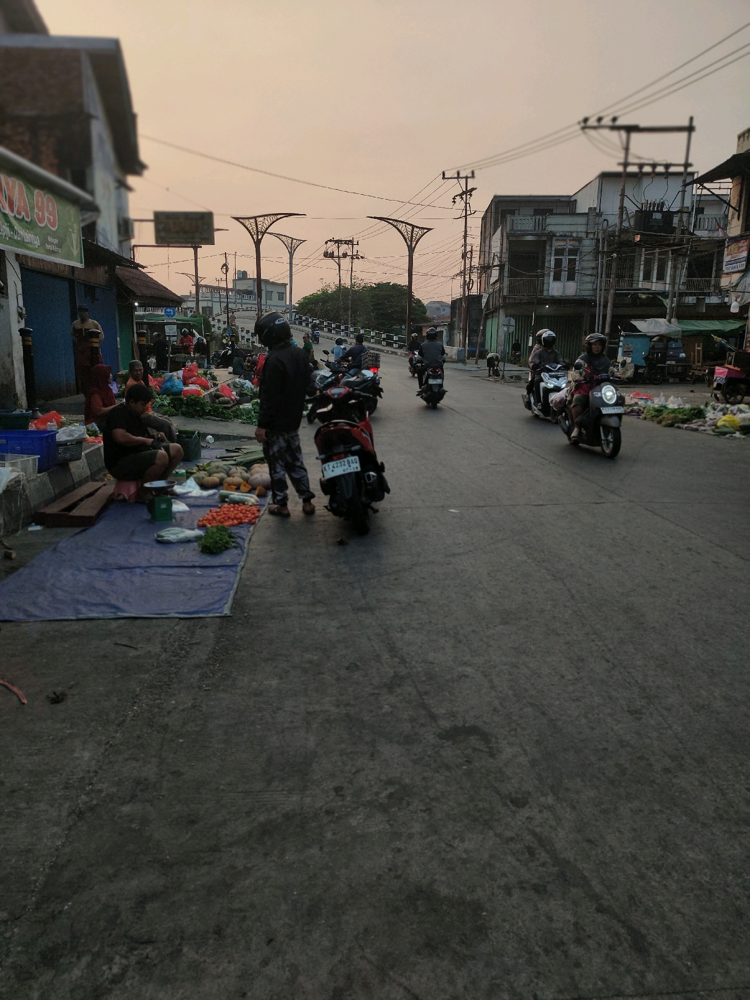 The image depicts a street scene in Samarinda, Indonesia, likely during the early morning or late afternoon given the soft, diffused light. The perspective is from ground level, looking down a paved street lined with buildings on the left and a market area extending onto the road on the right.
On the left side, a building with a green awning and the text "JAYA 99" is visible. Various goods, possibly produce, are laid out on blue tarps on the ground in front of some establishments. Several people are present in this area, some appearing to be vendors and others customers. A man in a black shirt and shorts is crouched down, possibly attending to the produce. Another person in a black jacket and patterned pants stands near a motorcycle.
The street itself is populated with numerous motorcycles, some parked and others in motion. Several individuals are riding motorcycles, wearing helmets. The road surface appears to be asphalt, with some debris and wear marks. Overhead, power lines and poles are prominent features against the hazy sky. The buildings on the right are multi-story structures, appearing somewhat aged, with balconies and exposed electrical wiring. Further down the street, more market stalls and motorcycles are visible. The overall impression is that of a busy, everyday street in a commercial or mixed-use area of Samarinda.