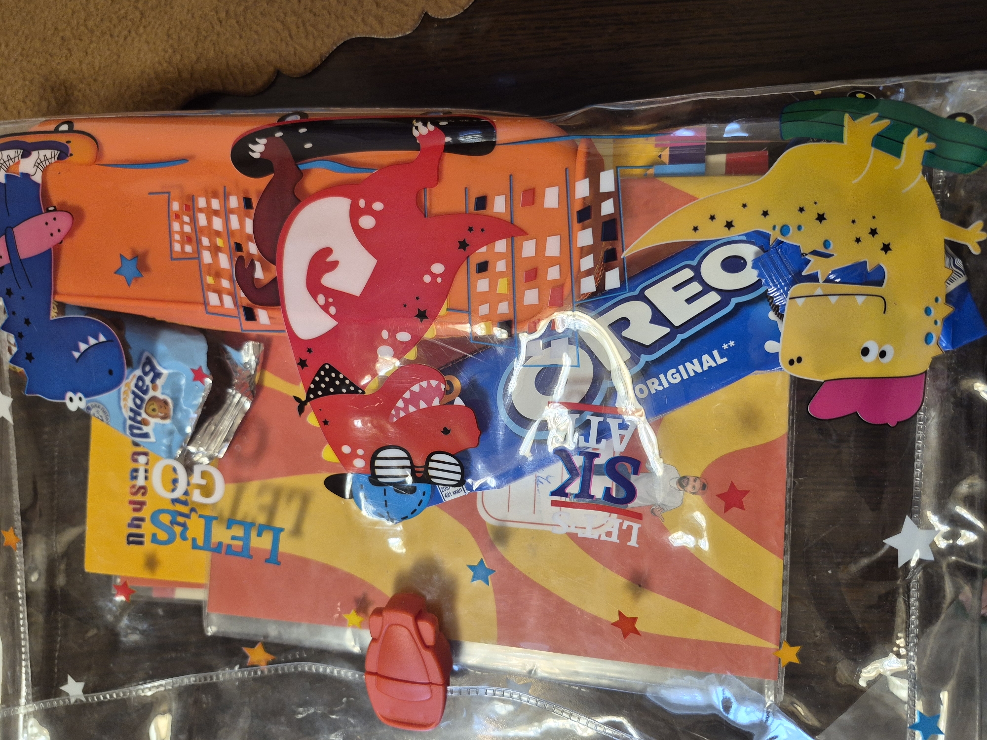 The image shows a clear plastic bag filled with various items, including a red toy, colorful paper cutouts of dinosaurs and buildings, and a package of Oreo cookies. The items are arranged in a visually appealing, organized manner. The background appears to be a wooden surface. There are no visible signs or landmarks that would indicate a specific location in Yerevan, Armenia. The overall scene suggests a collection of playful or decorative items, possibly intended for a gift or party.