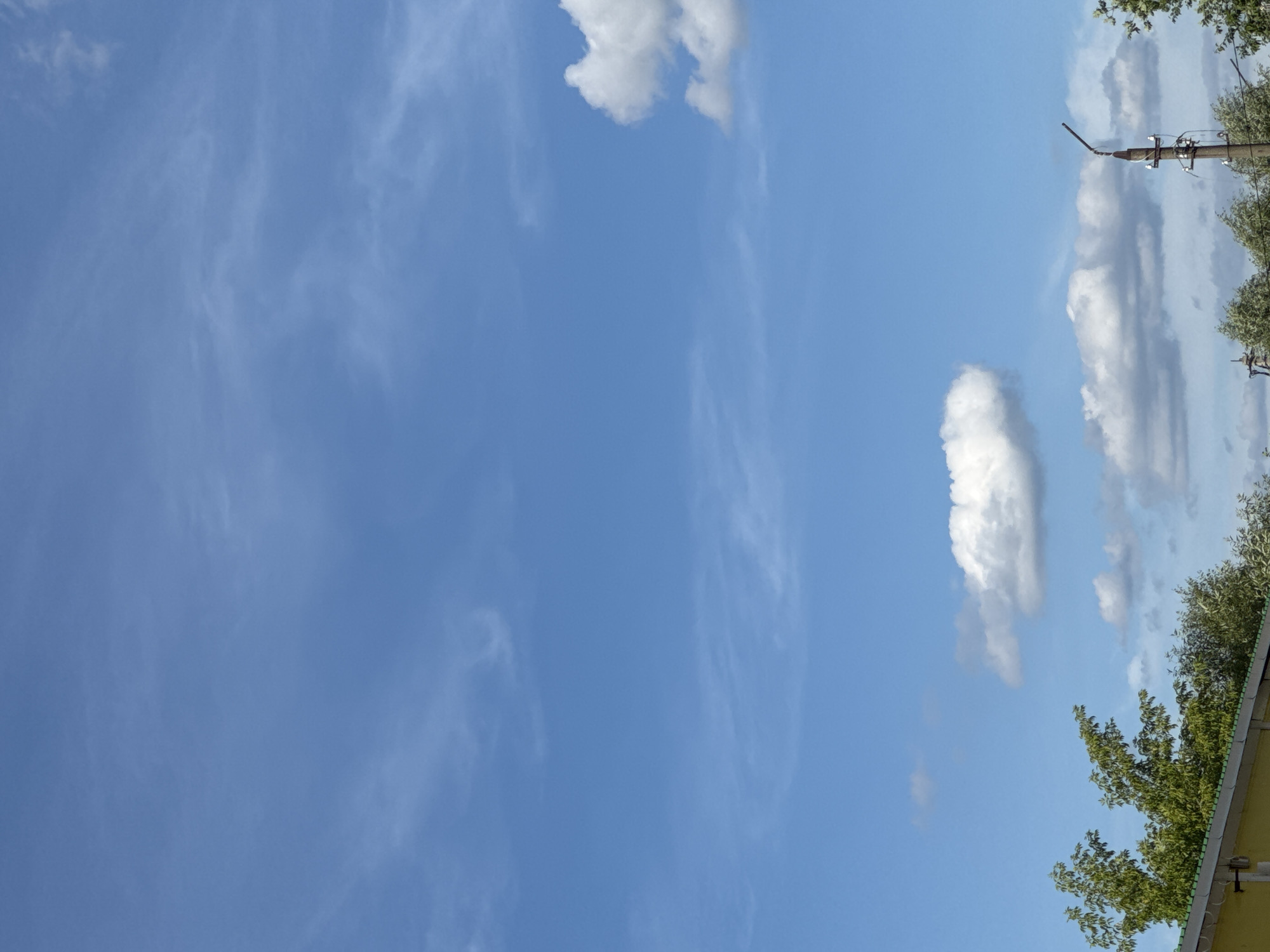 The image captures a clear blue sky with wispy white clouds, indicative of fair weather. In the lower portion of the frame, the tops of green trees are visible, along with the edge of a yellow building with a green roof. A utility pole with wires and a streetlamp is also present on the right side of the image. The scene appears to be an outdoor environment within a city, though no specific landmarks or identifying features place it in a particular location. There are no people or animals visible in the image.