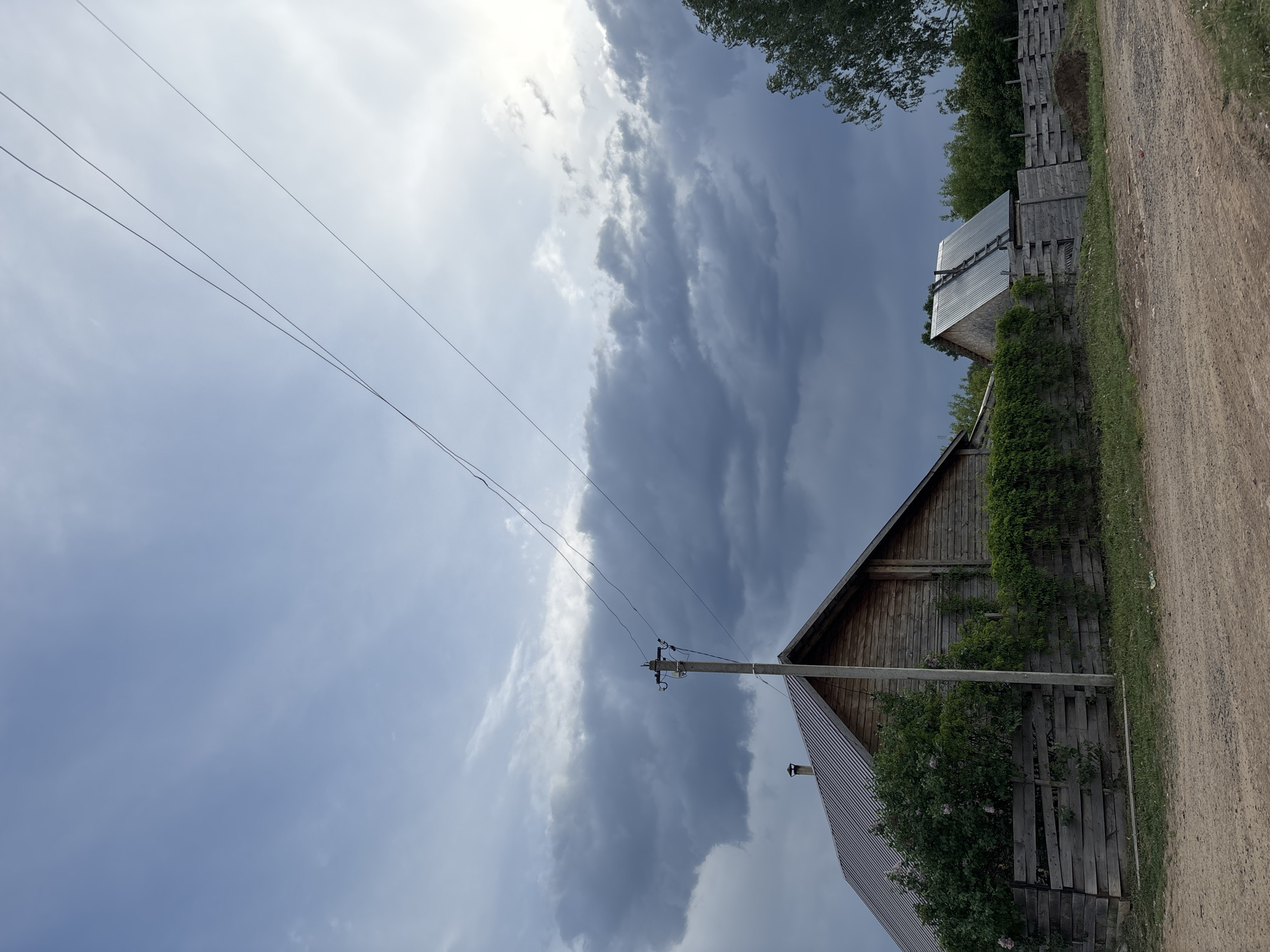 The image shows a rural scene in Фоки, Russia, featuring a dirt road alongside a weathered wooden fence partially obscured by greenery. Two wooden buildings with corrugated metal roofs are visible. An electrical pole stands beside one of the buildings, with three wires extending across the sky. The sky is filled with a mix of light and dark storm clouds. The photograph is taken from a low angle.