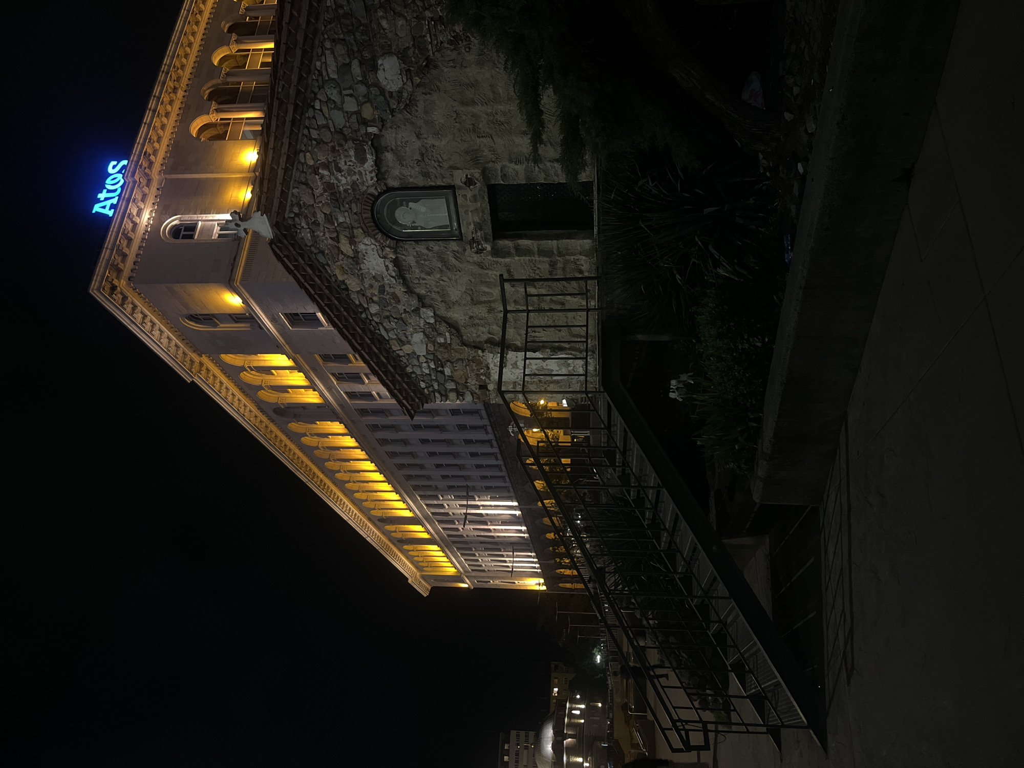 At night, the Atos Hotel in Sofia, Bulgaria, is captured from a low angle. The building's facade features illuminated arches and windows. A neon sign reading "Atos" is on the top. To the right, there's a section of stone wall with a framed image and a black opening. A metal staircase with black railings runs along the side of the building, leading down into a garden area with sparse plants. In the background, other buildings are visible with lit windows. A dark sidewalk occupies the right foreground.