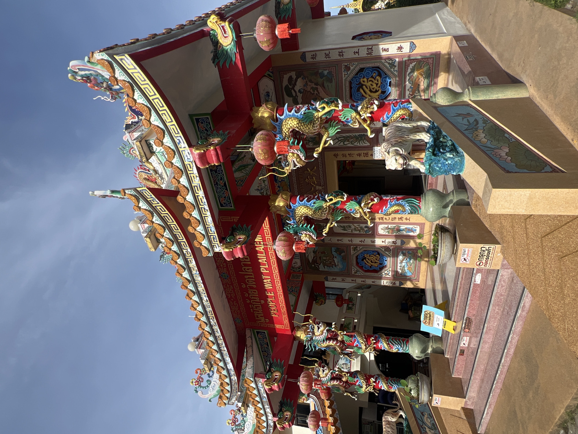 This is a photograph of the exterior of a temple in Baan Plai Laem, Thailand. The temple features traditional Chinese architectural elements, including a brightly colored, multi-tiered roof adorned with decorative figures and ornate tile work. The facade is painted red and white, with intricate murals and calligraphy on the walls. Numerous dragon sculptures are integrated into the structure, particularly around the entrance, with some appearing to coil around pillars. Red lanterns hang from the eaves. A white tiger statue stands on a pedestal near the entrance steps. The entrance itself is approached by a set of stairs. Signs are posted near the entrance, some bearing Thai text and images indicating that shoes should be removed. The sky above is clear and blue.