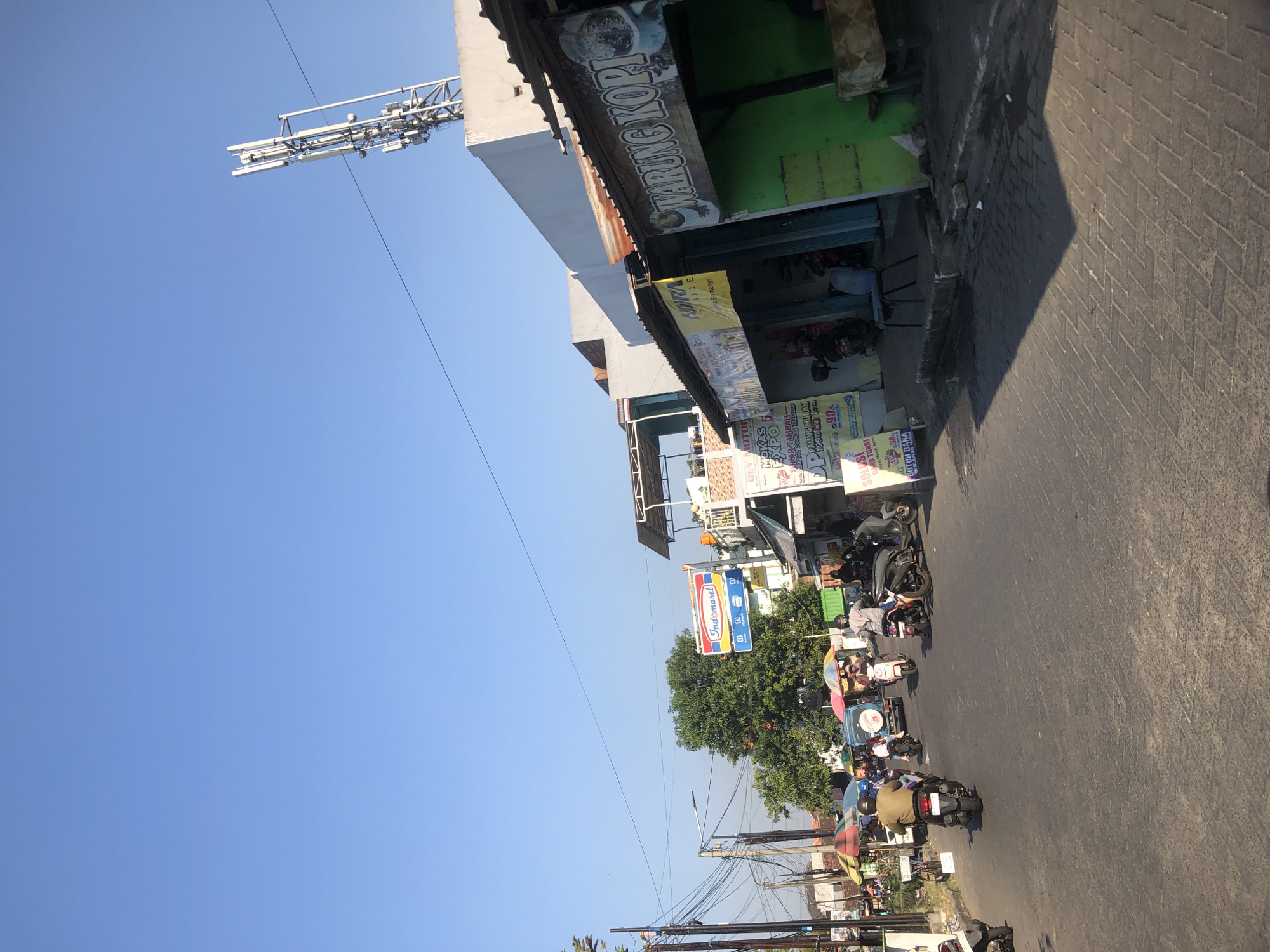 The image depicts a street scene in Boro, Indonesia, under a clear blue sky. The foreground is dominated by a paved road with textured bricks. Several motorcycles are present, with some individuals riding them and others parked. On the right side of the road, there are storefronts. One storefront features a green facade and a sign that reads "WARUNG KOPI" (coffee stall) with an image of coffee. Next to it, another storefront has a yellow awning and advertisements. Further down the street, on the left, there are more shops and stalls, including one with an "Indomaret" sign, a convenience store chain. A large green tree stands behind some of these shops. A telecommunications tower is visible in the upper right corner of the frame, situated on top of a white building. The overall impression is that of a busy, commercial street in a town or city.