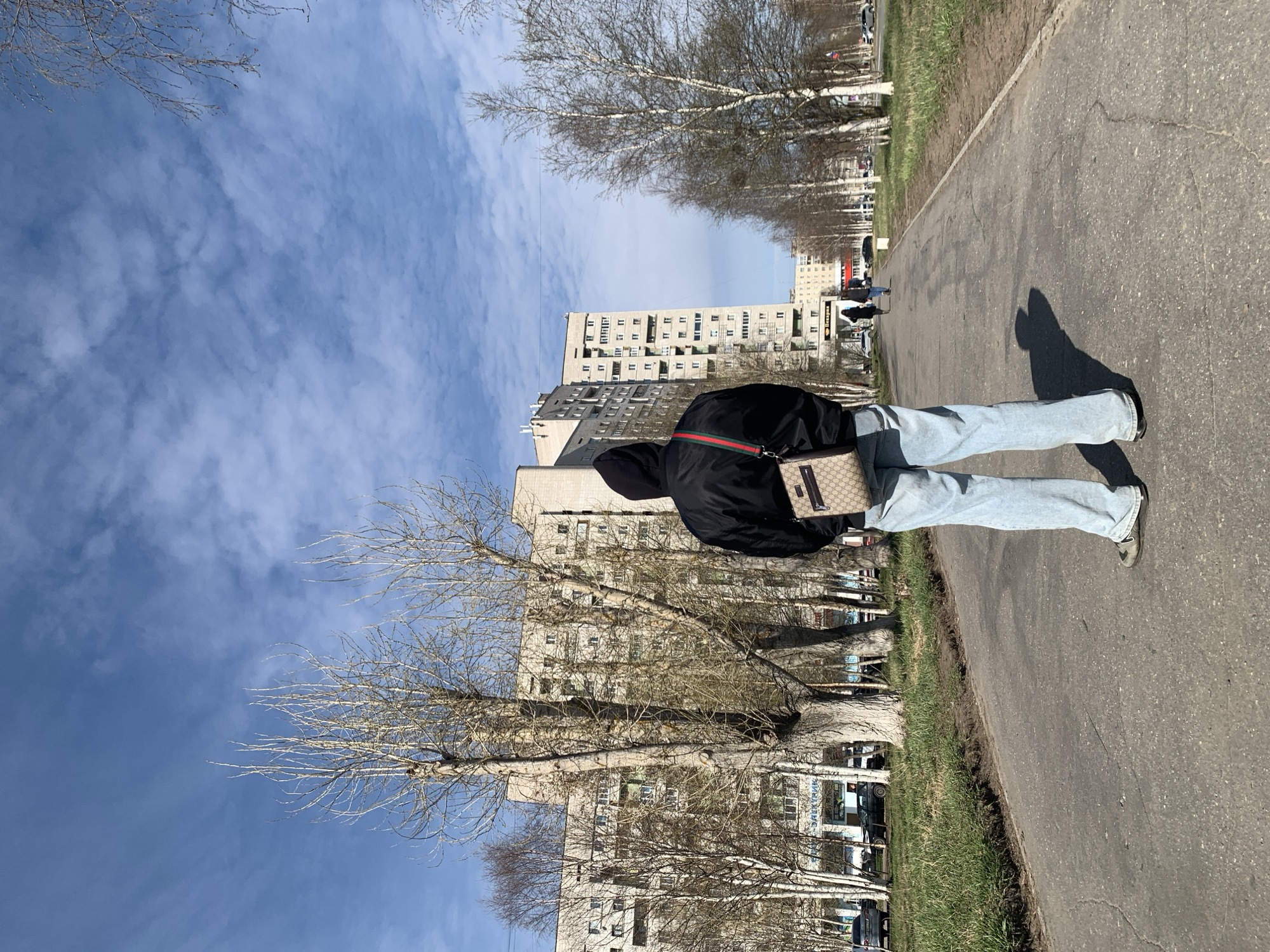 A medium shot, taken outdoors, shows a person walking on a paved path next to a road in what appears to be a residential area in Severodvinsk, Russia. The person is facing away from the camera, wearing a black jacket with a red and green stripe detail, light blue jeans, and grey shoes. They are also carrying a beige-colored shoulder bag. The path is made of grey asphalt, and to the right of the path is a strip of green grass. Parallel to the path and grass is a road with several parked cars. Across the road are numerous tall residential buildings made of concrete, with rows of windows. The sky is blue with scattered clouds, and there are bare trees lining the path. The light suggests it is daytime. The person's shadow stretches along the path in front of them.