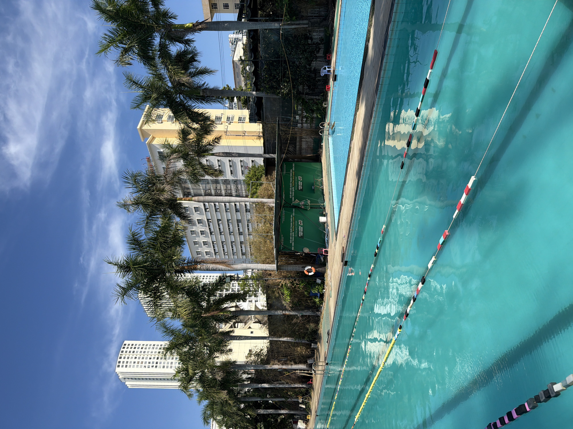 The image shows a swimming pool next to palm trees and tall buildings under a blue sky. The pool has lane dividers with red, white, black, and yellow markers. A life preserver hangs near the pool. Behind the pool, there are some green signs with Vietnamese text. The buildings indicate an urban setting, potentially in Nha Trang Ward, Vietnam.