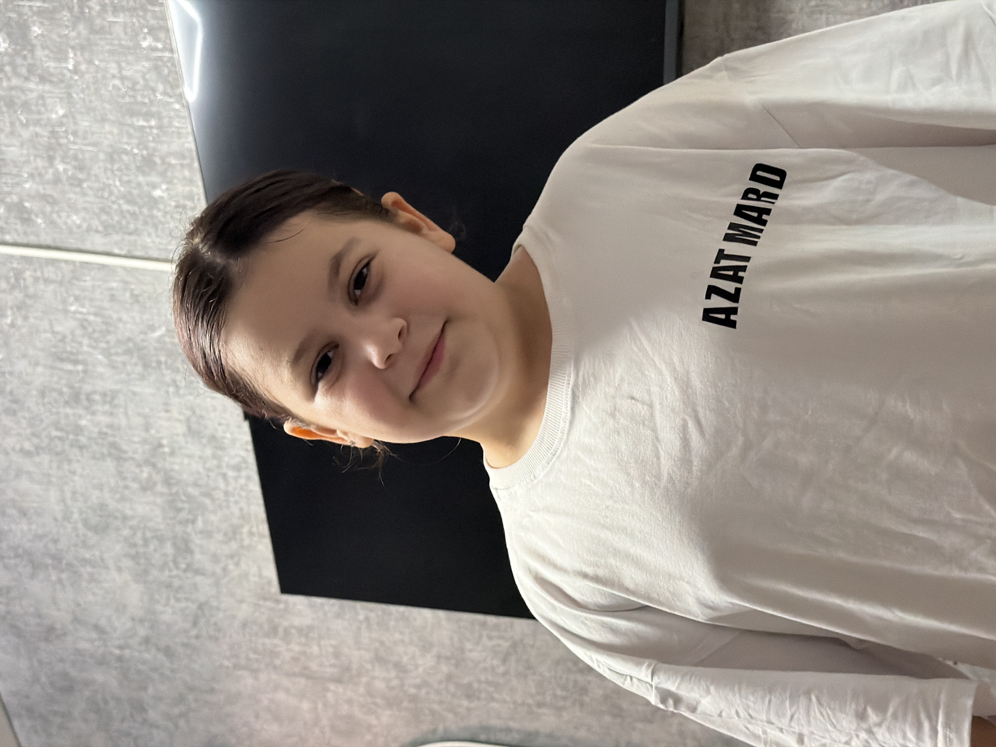 A young girl, likely a teenager, is the sole subject in this media file. She is wearing a white long-sleeved t-shirt with black text that reads "AZAT MARD" printed vertically on the upper right side of her chest. Her hair is dark and pulled back. The girl is looking at the camera with a slight smile, her head tilted to the side.
The background is dimly lit and appears to be an indoor setting. To the left, there's a wall with a textured, light grey surface, possibly concrete or stone. To the right, a large, dark, rectangular object dominates the background, potentially a screen or a piece of furniture. Above this, a bright horizontal light fixture is visible. The lighting suggests it could be daytime, but the indoor setting and lack of natural light cues make it difficult to determine the exact time of day or weather conditions. The overall mood is casual and informal, possibly a snapshot taken at home. No specific location cues beyond the general indoor setting are present, other than the provided context of Tuapse, Russia. The girl's expression conveys a sense of gentle amusement or contentment.