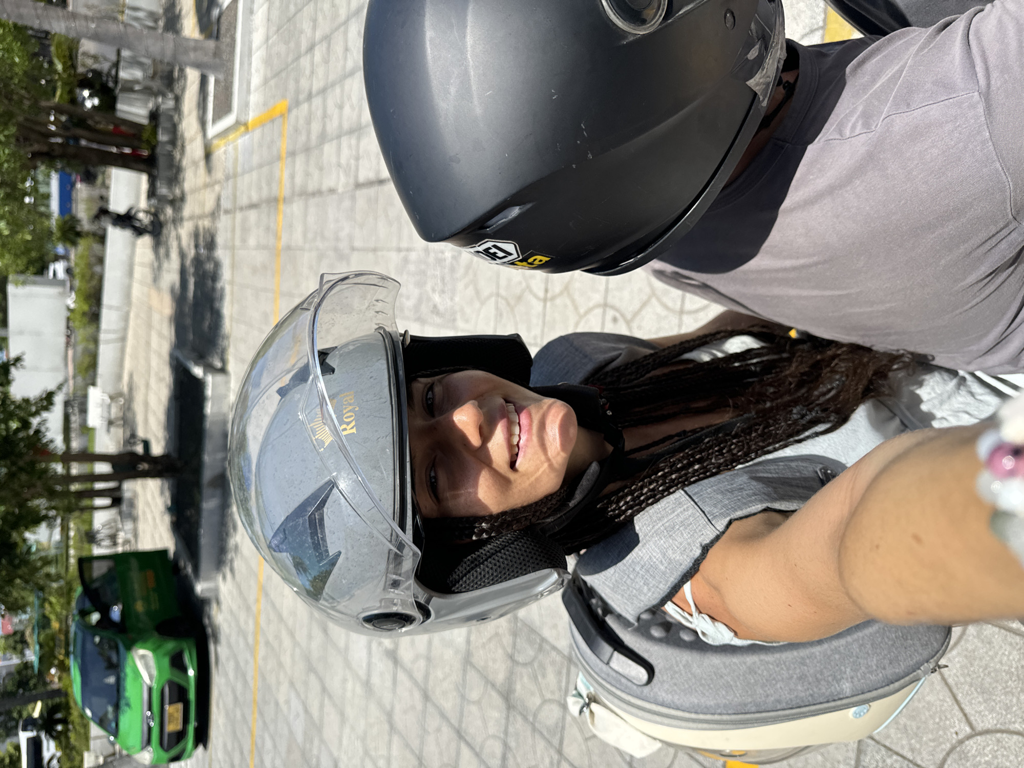 The image shows two people on a motorbike in Phu Thuy Ward, Vietnam. The person in the foreground is a woman wearing a silver helmet with a clear visor and a gray backpack. She is smiling and looking at the camera. Her hair is braided and falls down her back. The person behind her is wearing a black helmet. They are on a paved area with yellow lines painted on the ground. In the background, there is a green car parked, trees, and some white benches. The lighting suggests it is daytime.