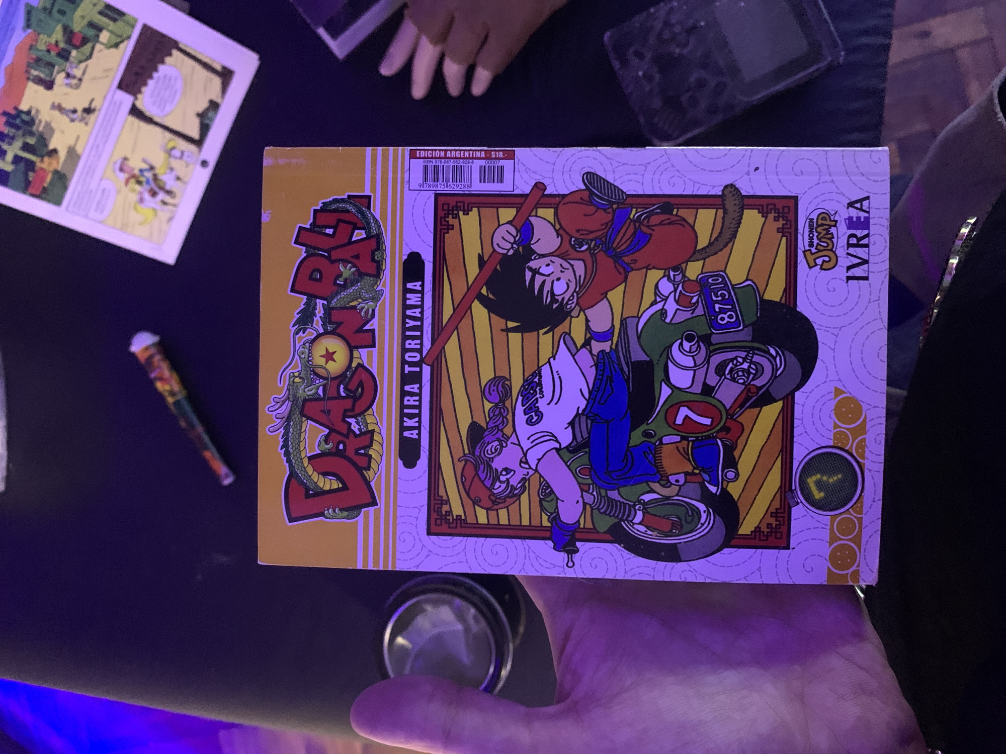 The image shows a close-up of a "Dragon Ball" manga book, specifically "Dragon Ball 7" by Akira Toriyama. The cover art depicts characters Goku and Bulma on a motorcycle. Text on the cover identifies it as "Edición Argentina - $18". The publisher is Shonen Jump IVREA. A hand is holding the book. Other items are partially visible in the background: a black object with buttons, a colorful pen, and a page from another book or magazine. There is no indication of the city of Buenos Aires.