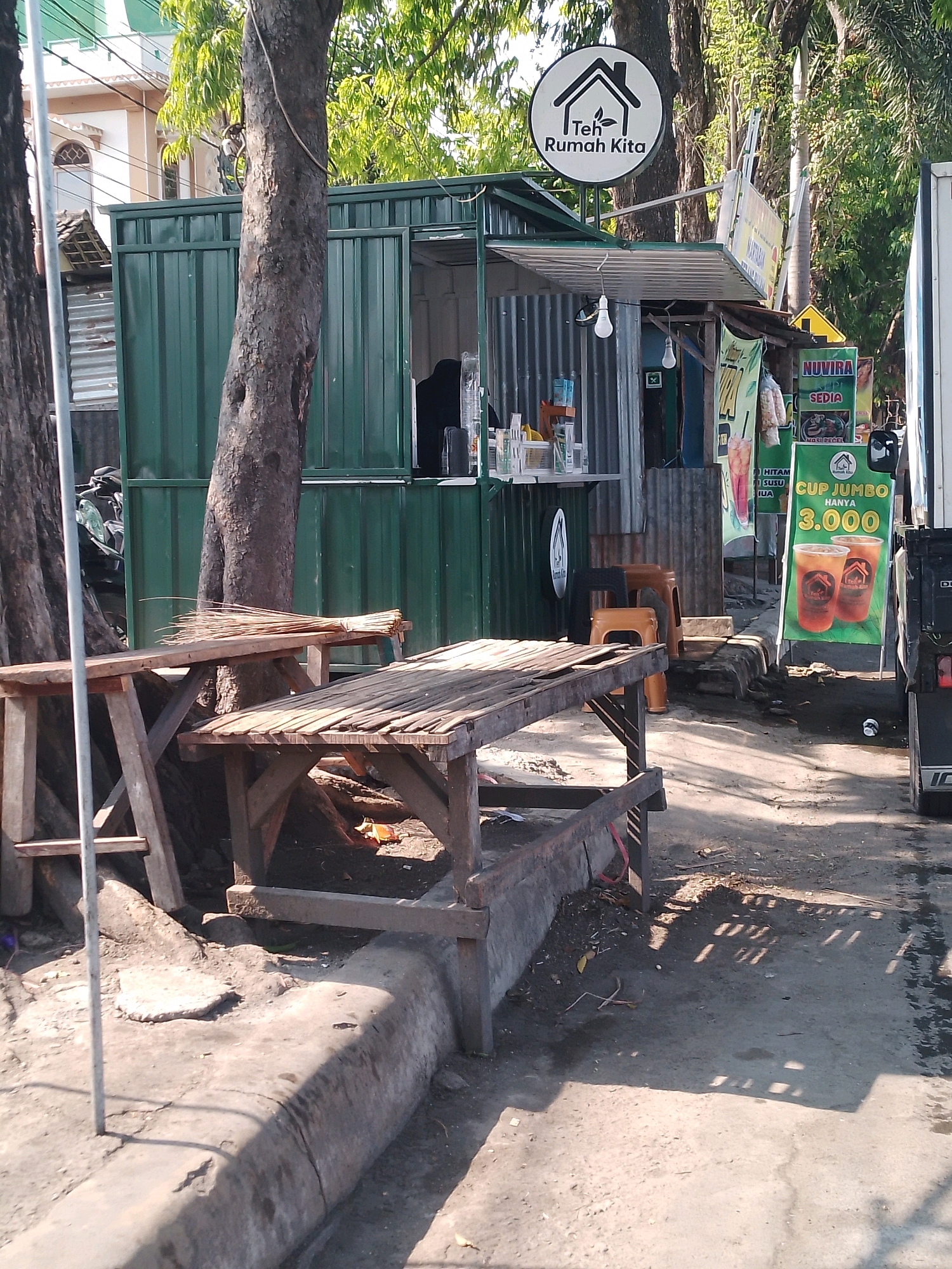 The image depicts an outdoor scene in Kertosono, Indonesia, likely a street-side food stall. A green corrugated metal kiosk with a white awning serves as the main structure. A circular sign with a house and leaf emblem above the text "Teh Rumah Kita" is prominently displayed. Various banners with text and images of drinks are hung on the kiosk and nearby poles. In front of the stall, two weathered wooden tables and benches are set up on a concrete curb. A bundle of dried palm fronds, possibly a broom, rests on one of the tables. The ground is a mix of asphalt and dirt, with shadows cast by trees and the structures. In the background, a building with a white facade and arched windows can be seen. A motorcycle is parked to the left of the kiosk. A truck is partially visible on the right side of the frame. The overall environment suggests a casual, everyday setting, typical of a local business in Indonesia.