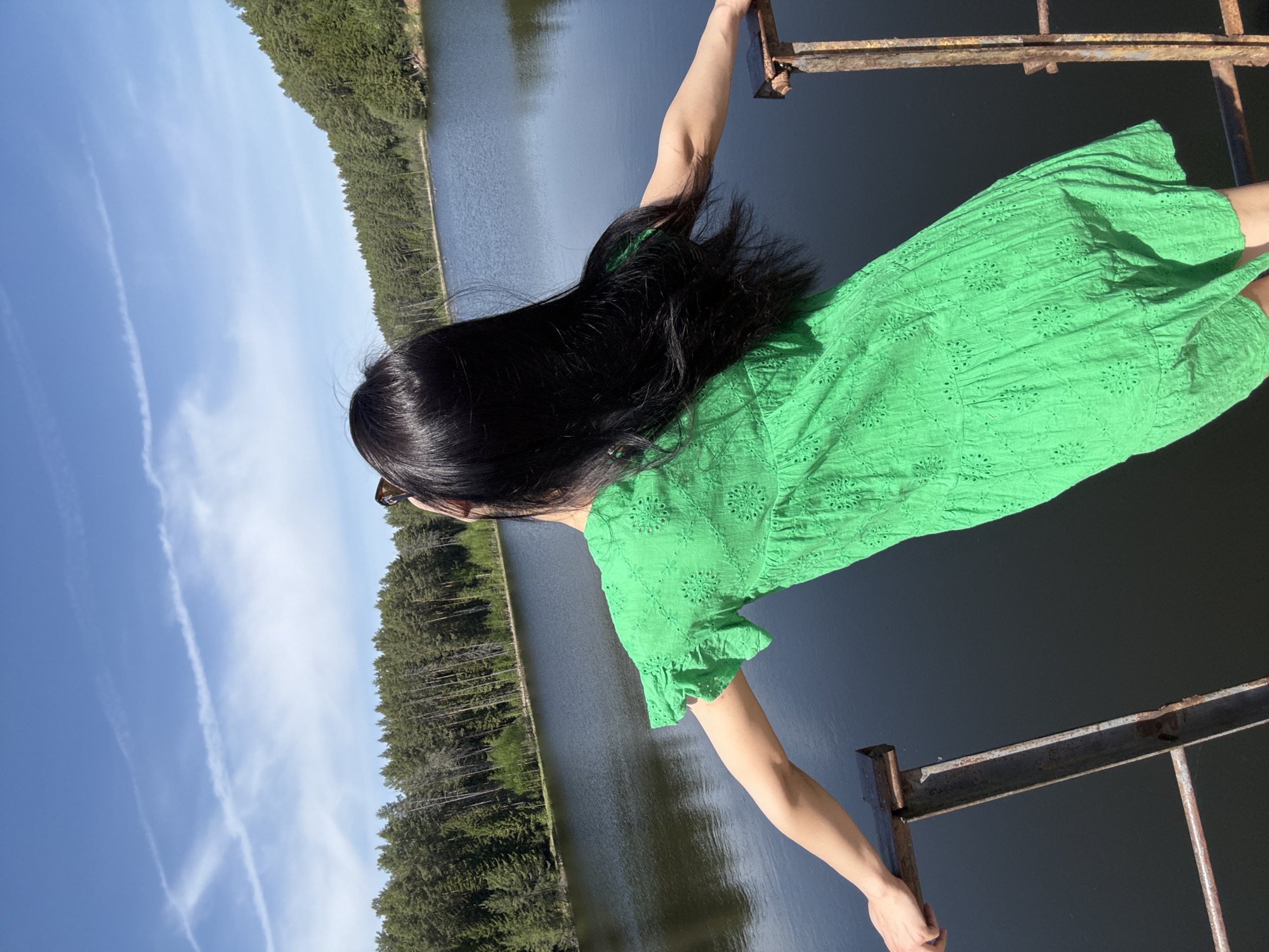 A woman with long, black hair stands on a rusty metal structure overlooking a body of water, likely a reservoir or lake. She is wearing a bright green dress. The backdrop consists of a dense forest line on the opposite side of the water and a blue sky with some cloud formations. The overall composition and environment suggest a location near a natural body of water, possibly in a recreational or scenic area.
Given the user prompt regarding Votkinsk, Russia, it is highly probable that the body of water is the Votkinsk Reservoir, and the photograph may be taken from a bridge or similar structure overlooking it.