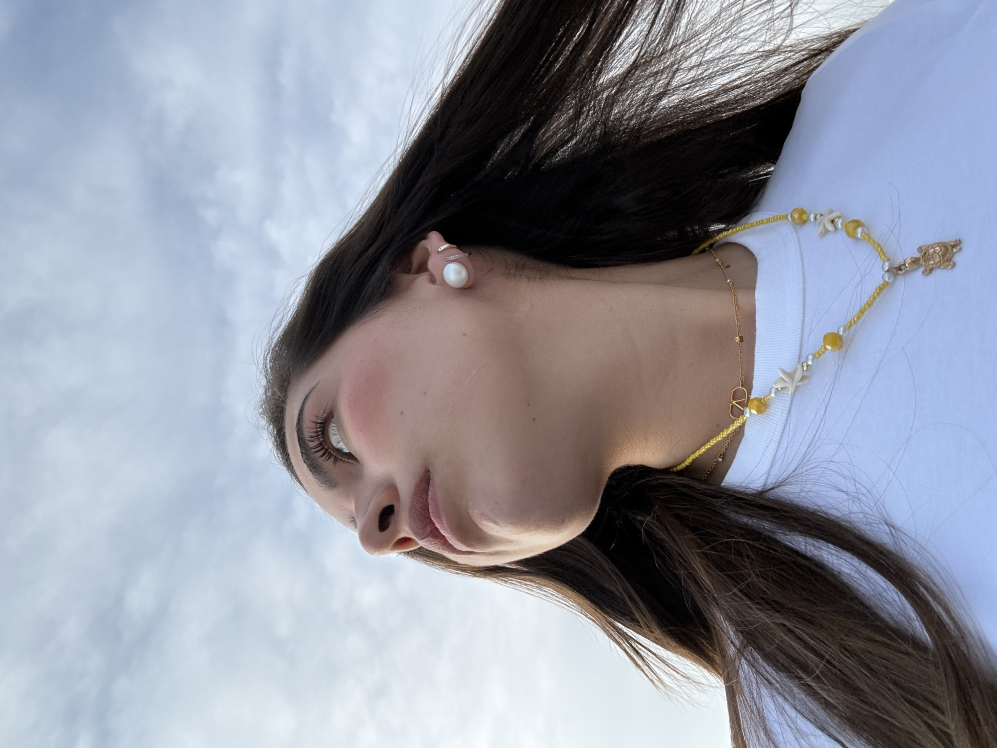 A medium close-up photograph shows a young woman from a low-angle perspective against a cloudy sky. The camera angle is rotated counter-clockwise. The woman has dark, wavy hair, and her face is visible in profile. She wears a pearl earring. A yellow beaded necklace with starfish charms and a turtle pendant is visible around her neck. She is wearing a white shirt. The image provides no information about the location of San Bernardino Tlaxcalancingo, Mexico.