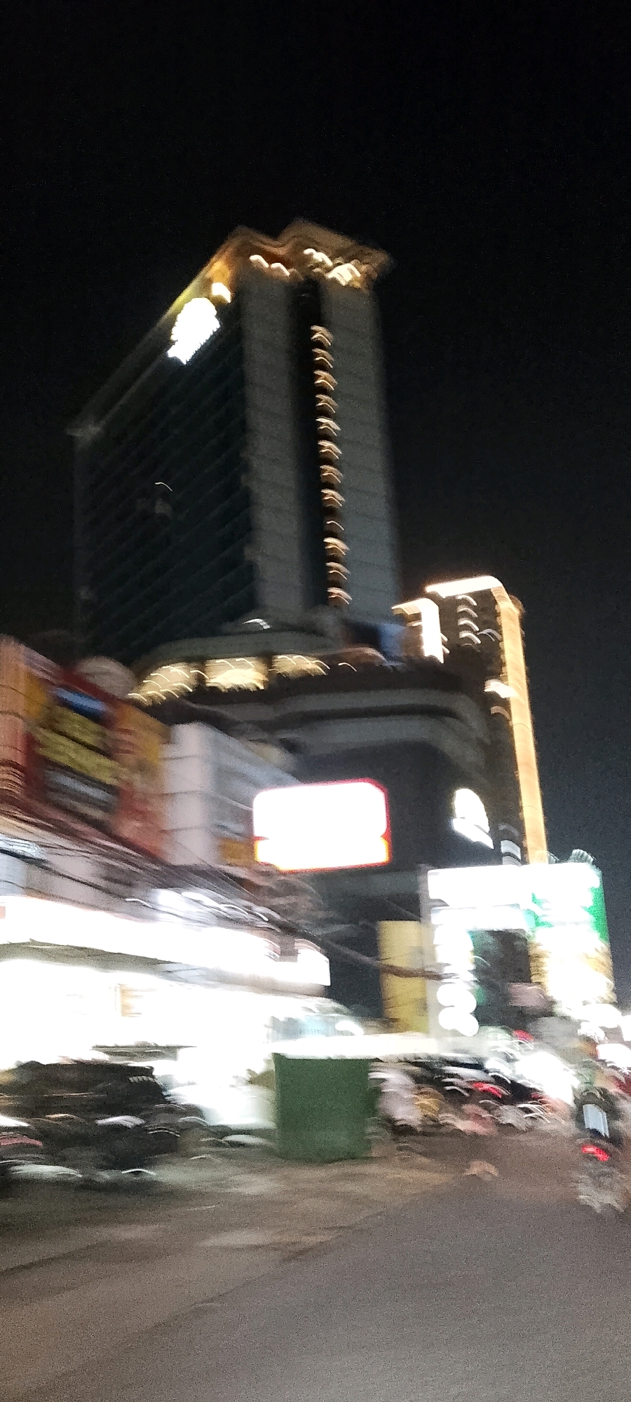 The image depicts a nighttime street scene in Depok, Indonesia, characterized by motion blur from a moving vehicle. Dominating the background are two brightly lit modern buildings, the taller one with a distinctive illuminated logo at its apex. Below these structures, commercial storefronts are visible, adorned with neon signs and various displays. In the foreground, the blurred forms of vehicles and a green dumpster suggest urban infrastructure. The overall impression is a dynamic urban environment at night, viewed from a moving perspective.