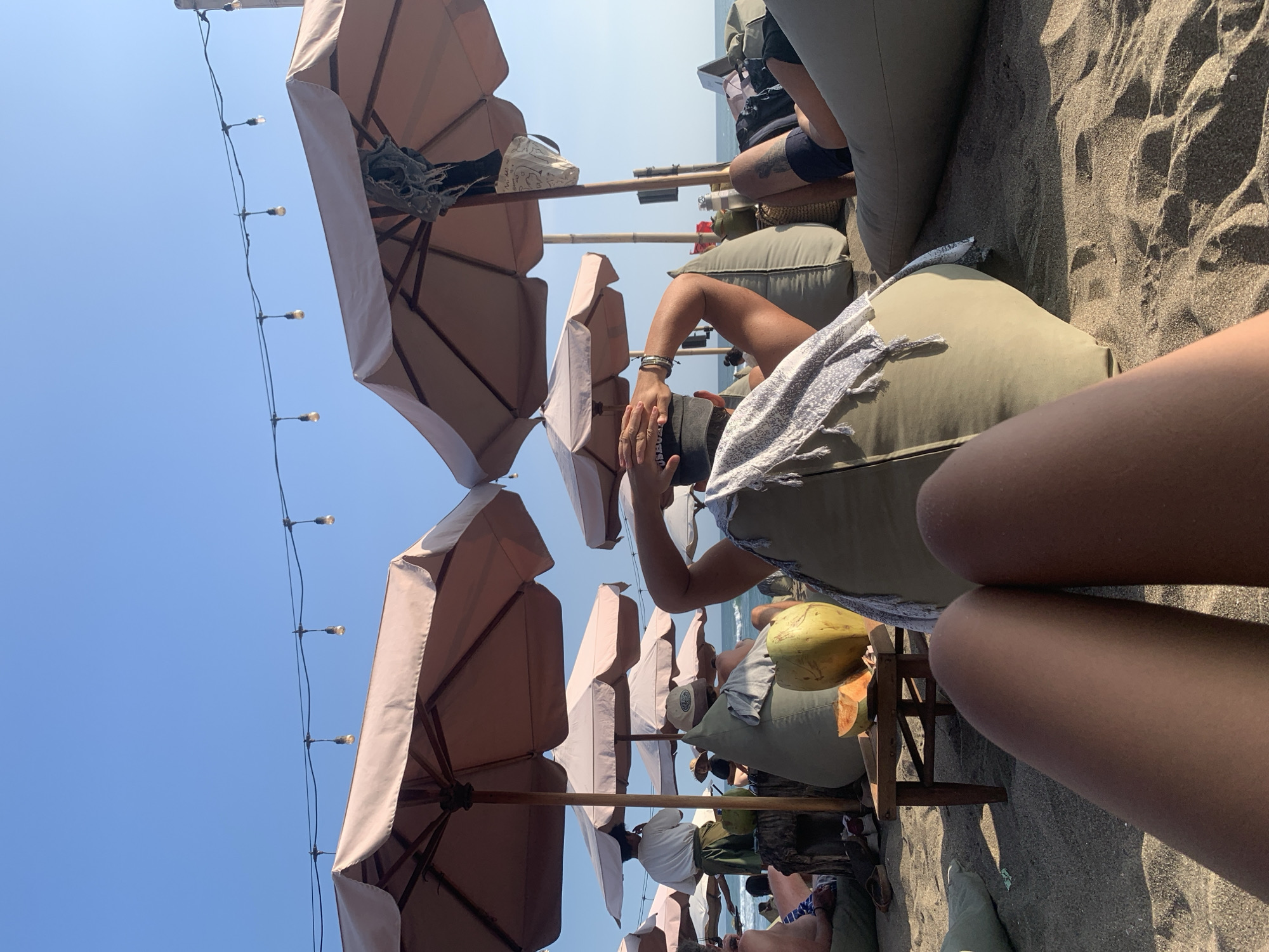 The image depicts a beach scene in North Kuta, Indonesia, with a clear blue sky. In the foreground, a person's legs are visible, resting on the sand. Behind them, several large, light-colored beach umbrellas provide shade over beanbag chairs. A small wooden table is placed in front of some of these chairs, holding a cut melon and a whole coconut. In the middle ground, more beanbag chairs and people are scattered, some seated and others appearing to be lounging. A waiter in a white shirt and green pants is visible in the background, interacting with patrons under an umbrella. String lights are visible overhead, stretching between poles. The overall atmosphere suggests a relaxed beach club or restaurant setting.