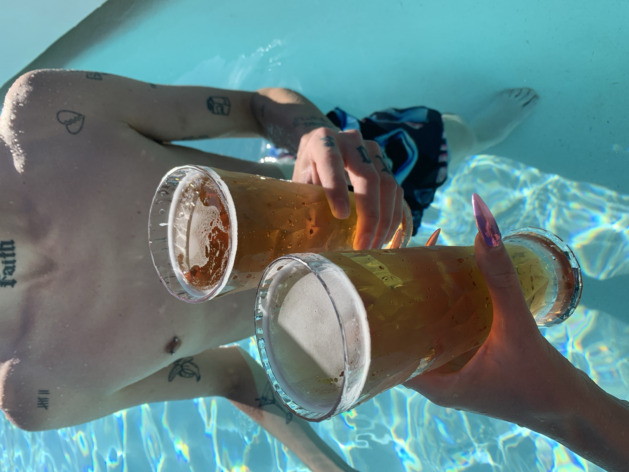 In an outdoor swimming pool under bright sunlight, two people are holding large beer glasses. One person is floating in the pool with a bare torso and tattoos visible on their upper body, arm, and chest. They are wearing dark swim trunks. The second person, visible only by their arm and hand holding one of the glasses, has long, metallic pink fingernails. The pool water is clear and reflects patterns of sunlight. The beer glasses are filled with a light-colored beer and have a foamy head. The scene appears relaxed and social. Olhão, Portugal, is not discernible from the image.