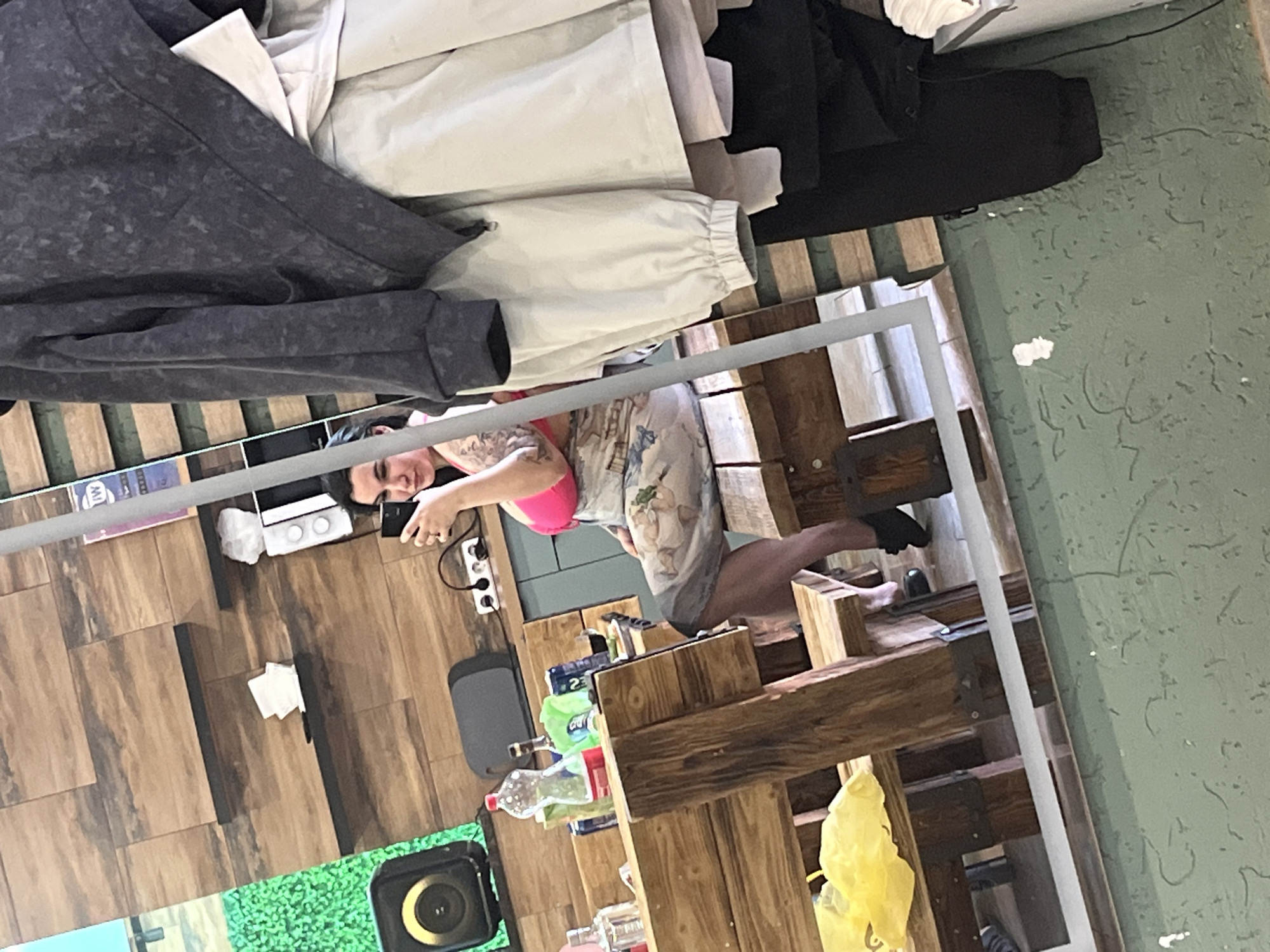 A mirrored image of a person using a phone is visible. The person is wearing a pink top and a patterned dress. They are standing in front of wooden structures, possibly shelves or a counter. The reflection shows a tiled floor, and various objects, including bottles and a speaker. Clothing items are also present on a surface above. The location is likely Chaikovsky, Russia, but there is no explicit confirmation in the image.