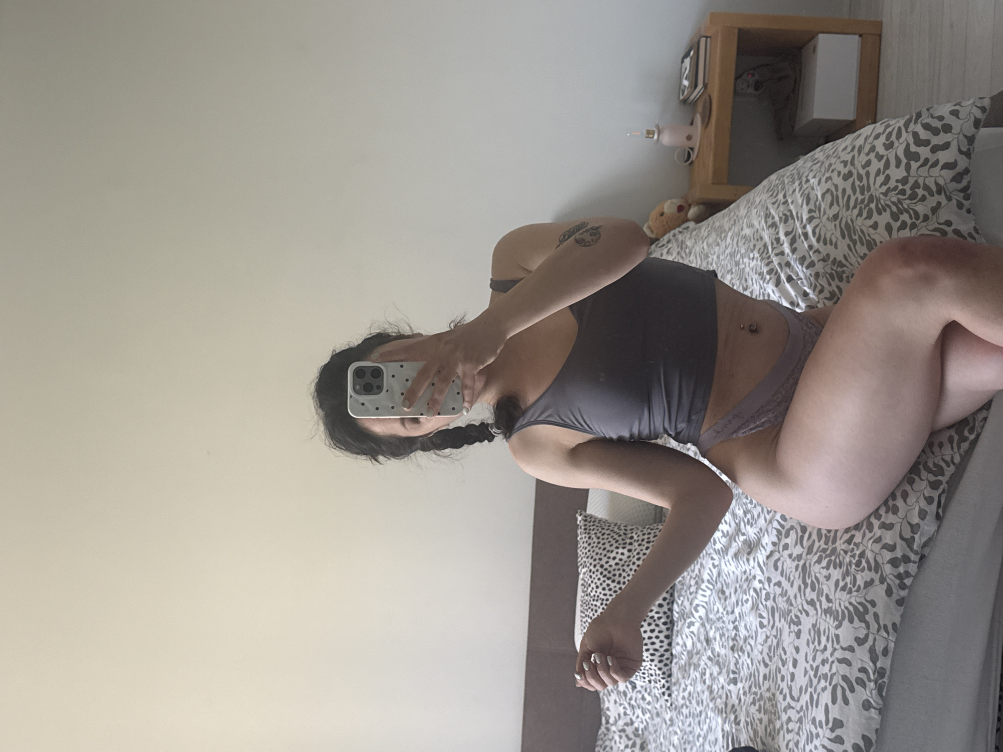 A woman in underwear and a top is lying on a bed with patterned sheets in a room. The woman is holding a smartphone in front of her face, seemingly taking a selfie in a mirror. Her hair is in pigtails, and a tattoo is visible on her arm. A nightstand with various items is next to the bed. The location is not discernible from the image, and there is no indication it is in Burgas, Bulgaria.