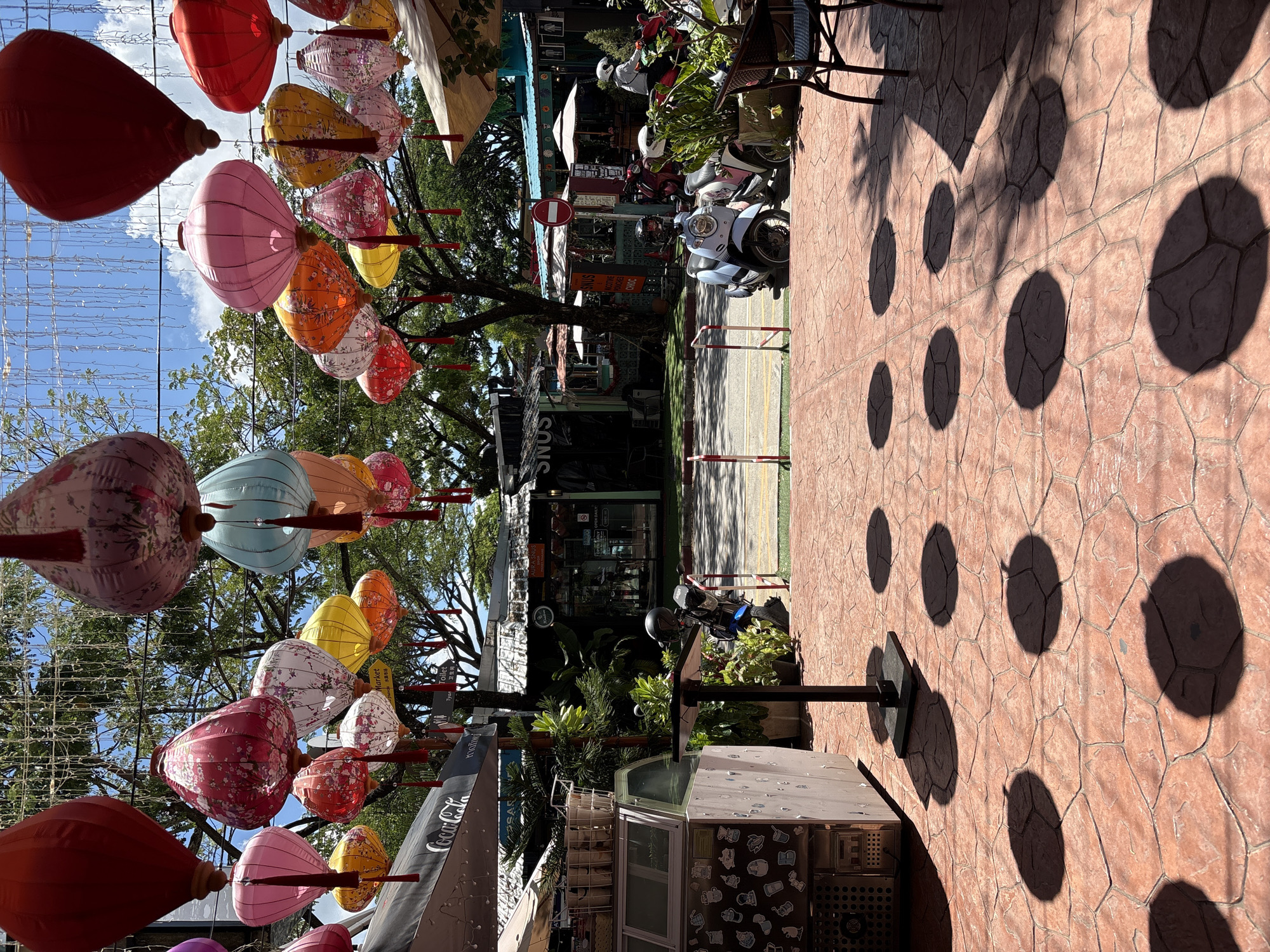 In Choeng Thale, Thailand, an outdoor marketplace is depicted under a bright, sunny sky. Colorful Chinese lanterns, in shades of red, pink, yellow, and blue, are suspended overhead, casting circular shadows on the terracotta-tiled ground. A light-colored kiosk with a glass display case is positioned to the left, adorned with numerous stickers. A dark-colored table with a scooter helmet on it stands in front of the kiosk.
In the background, several stalls and shops with pitched roofs are visible. A Coca-Cola umbrella shades a portion of the scene. Scooters are parked along the road, with one light blue scooter in the foreground and several others further back, including one being ridden by a person wearing a white helmet and shirt. A person in a white helmet and shirt is also visible riding a scooter in the distance. A few potted plants are placed near the scooters, and a chair with a table is situated on the right side of the foreground. The overall environment suggests a bustling, open-air market setting.