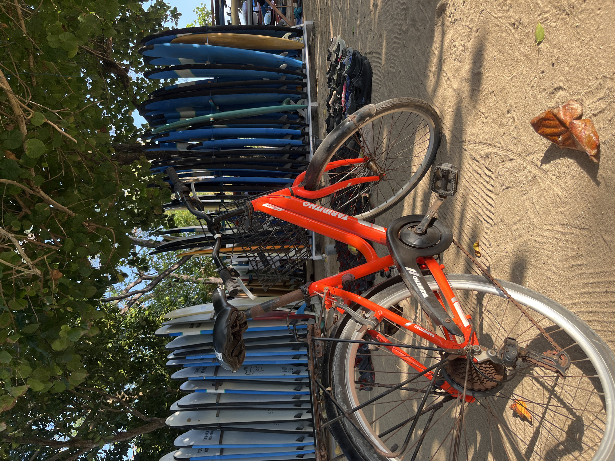 A red bicycle is parked on a sandy beach in Kuta Selatan, Indonesia, beneath the shade of trees. The bicycle has a basket on the handlebars and a rack over the rear wheel, with a rusty chain and gears visible. In the background, a row of surfboards stands upright, and a collection of footwear is neatly arranged on the sand. The overall scene depicts a relaxed atmosphere often associated with beachside locations.