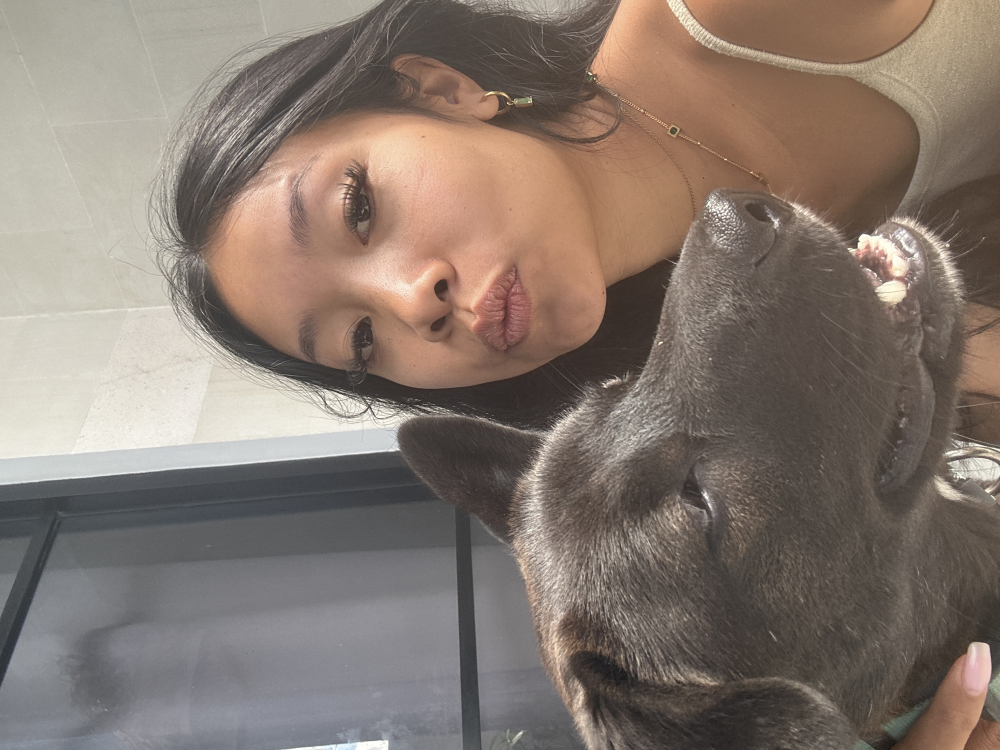 A young woman with long, dark hair and false eyelashes puckers her lips for a selfie. Beside her, a dark-furred dog's head is visible, its mouth slightly open, revealing its teeth. The woman wears a light-colored, sleeveless top and delicate gold necklaces, one with a green pendant. She also has a gold hoop earring with a green stone in her left ear. The background features a light-colored tiled wall and a dark-framed window or door. The scene appears to be indoors, possibly in a bathroom or a modern residential setting, with soft natural lighting. The location is not specified as being in Sukawati, Indonesia, or any other specific city.