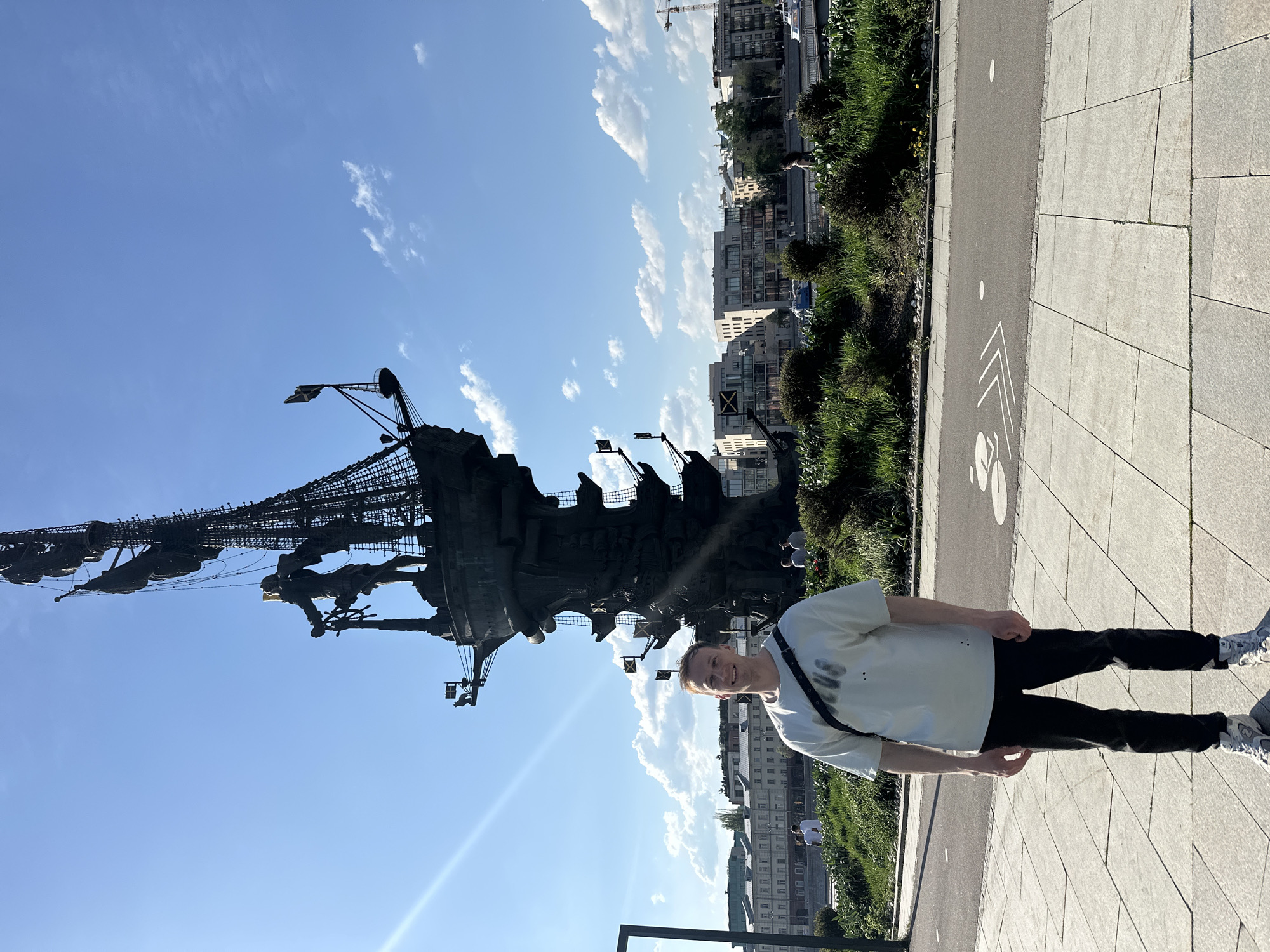 A vertically-oriented, full shot shows a man standing on a paved walkway next to a tall, dark-colored sculpture against a backdrop of buildings, green vegetation, and a blue sky with clouds. The man, positioned to the right, wears a light-colored t-shirt, dark pants, and light-colored sneakers. He has a dark-colored bag strapped across his chest. He is smiling and appears to be posing for the picture. To the left, the sculpture dominates the scene, and it seems to depict a large ship or boat made of metal. The sculpture is detailed with what appears to be figures of people. Behind the sculpture, buildings of varying heights are visible with trees and green spaces interspersed. The paved walkway is made of light grey stone blocks with a designated lane for cyclists. The sky is clear blue with scattered white clouds, and a lens flare is visible. This scene is in Moscow, Russia.