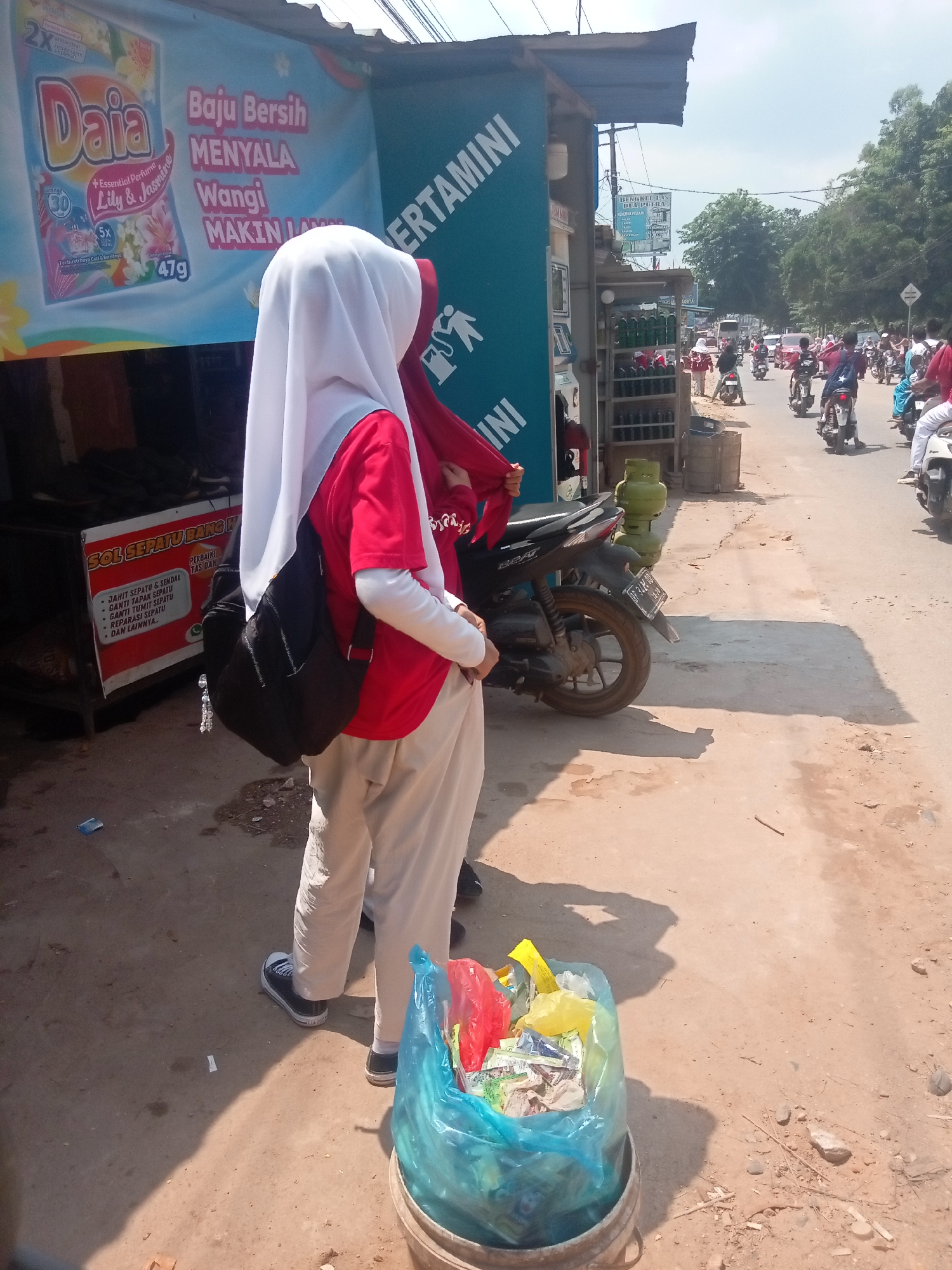 In Batam, Indonesia, on a sun-drenched street with a cracked asphalt surface and patches of exposed dirt, a scene unfolds near a roadside stall. Two individuals, likely students based on their attire, stand in the foreground. The person on the left is wearing a white hijab and a red long-sleeved shirt with cream-colored pants, carrying a black backpack. The person to their right is partially visible, wearing a red hijab.
Dominating the background is a large banner advertising "Daia" detergent, featuring a product image and Indonesian text emphasizing clean, fragrant, and long-lasting clothing. Adjacent to this, a blue sign with white text reads "ERATAMINI." To the right of the banner, a stall displays an assortment of bottled beverages. Further down the street, a procession of motorcycles and a few cars navigate the road, indicating a moderately busy thoroughfare. A black scooter is parked in the middle ground.
In the immediate foreground, a bucket filled with various colored plastic bags holds discarded waste, including red, yellow, and blue bags, with visible trash within them. The overall environment suggests a commercial area with a blend of advertising, local businesses, and daily commuter activity. The lighting suggests it is daytime, likely mid-morning or afternoon, given the strong shadows cast by the individuals and objects.