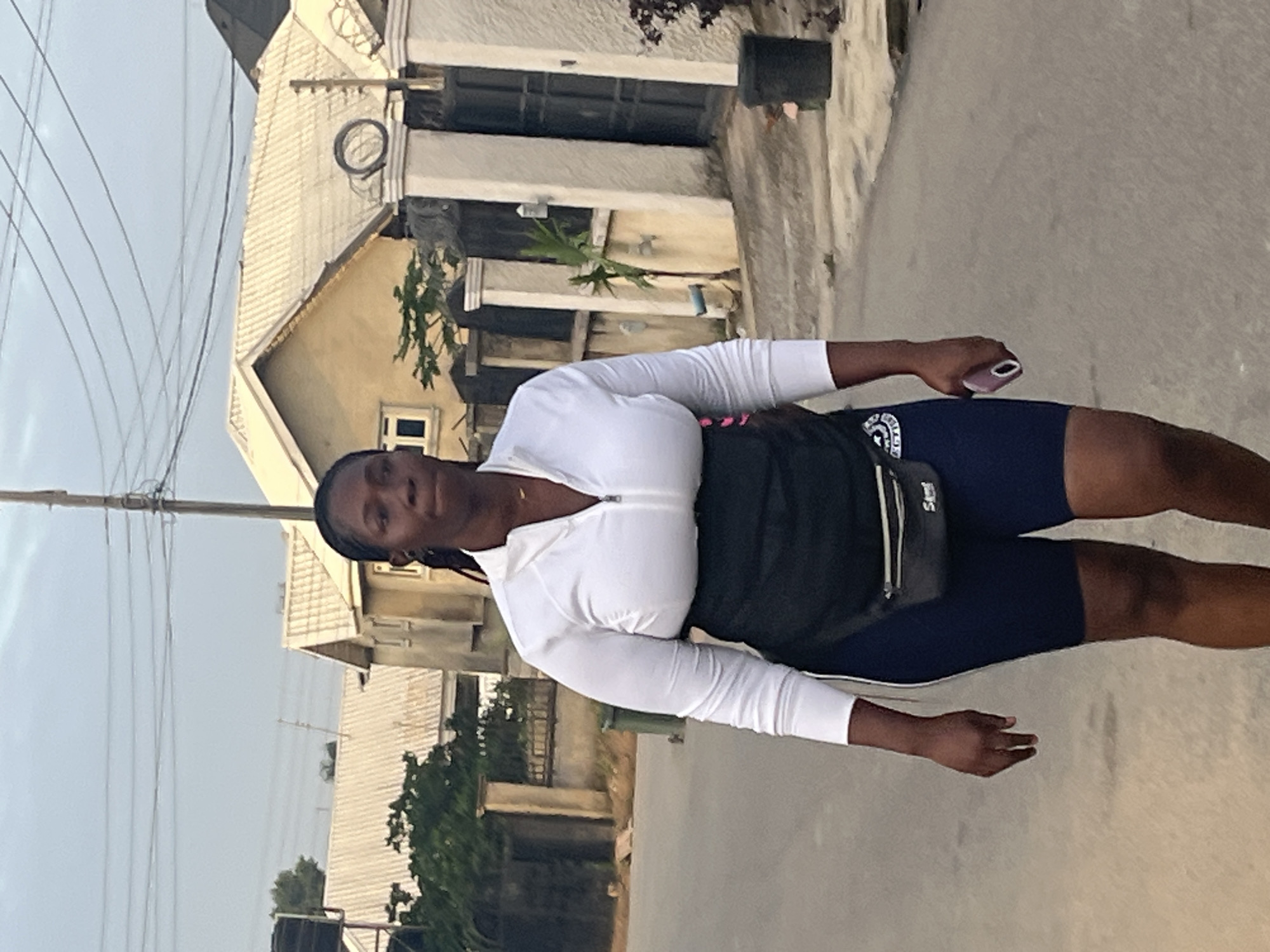 A dark-skinned woman with braided hair is walking on a paved street in Abuja, Nigeria. She is wearing a white zip-up long-sleeved shirt, dark blue athletic shorts, and a black fanny pack around her waist. She is holding a light pink cell phone in her right hand. The background shows residential buildings with corrugated iron roofs and some trees. The sky is overcast.