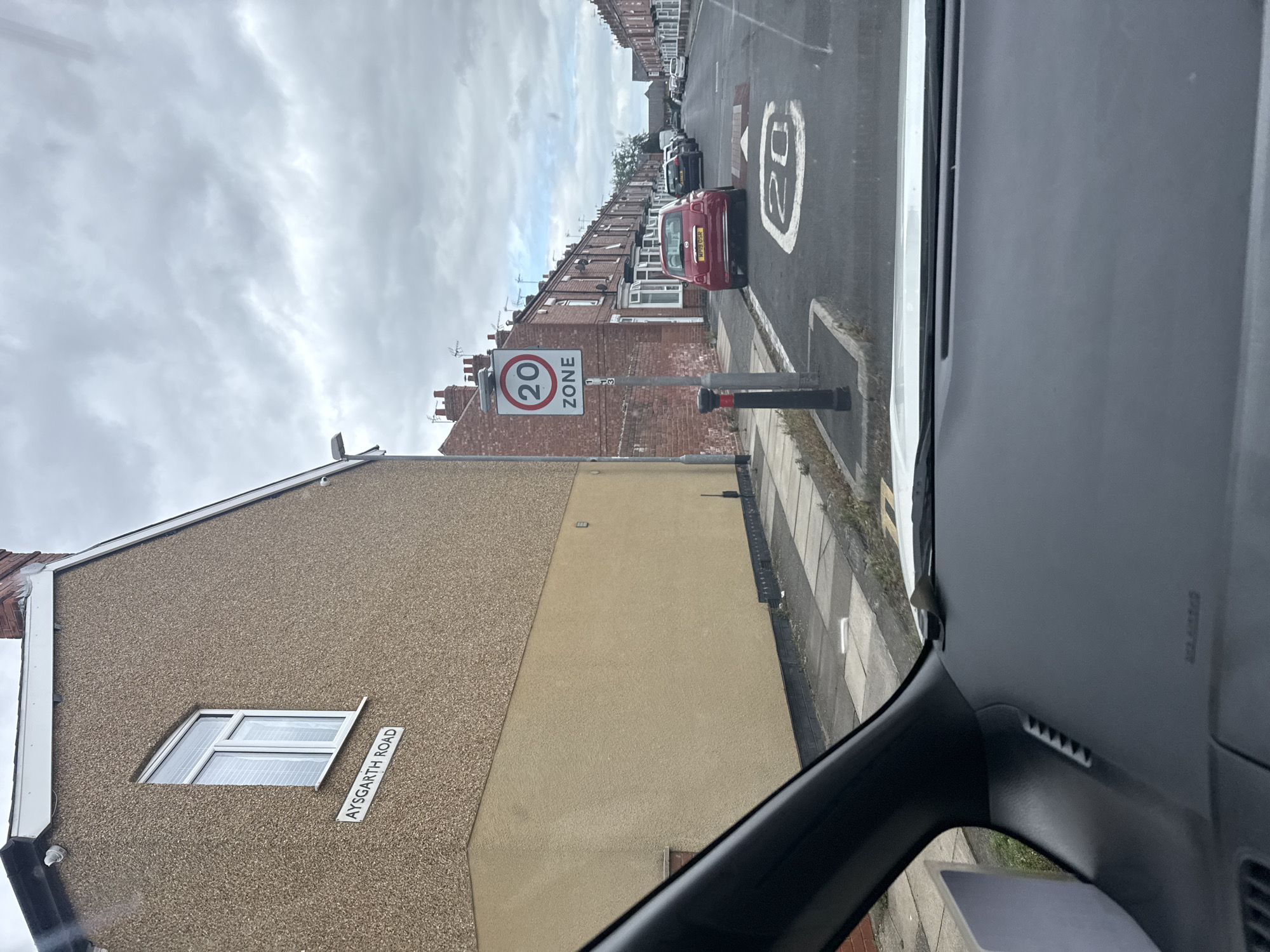 This image captures a street scene in Darlington, United Kingdom, likely during the daytime on a cloudy day. The perspective suggests the photo was taken from inside a vehicle. The foreground features a textured, light brown wall of a building with a distinctive angled window and a street sign that reads "AYSGARTH ROAD".

The middle ground shows a street with parked cars and a red hatchback. A traffic sign indicates a "20 ZONE". The street is lined with terraced houses made of red brick, with chimneys visible on their roofs. The pavements are paved, and there is some greenery along the side of the road.

In the background, the street continues with more terraced houses and cars, receding into the distance. The sky is overcast with a pattern of grey clouds. The overall impression is of a residential street in a built-up area. There are no people visible in the image.