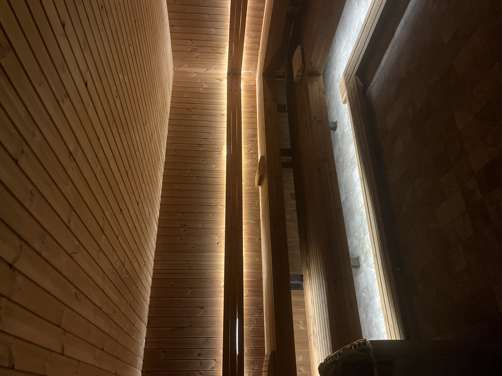 The image shows the interior of a sauna, possibly located in Sofia, Bulgaria. The walls are covered in horizontal wooden planks, some with linear lighting strips. Vertical wooden beams are visible. A stone or tiled surface is seen in the background, and an object is partially visible in the foreground, possibly a sauna bench.