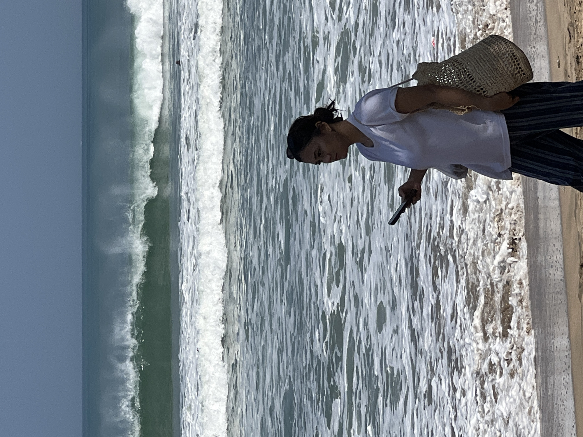 A woman walks on the beach in Kuta, Indonesia, with waves crashing in the background. She is wearing a white t-shirt and striped pants, and is carrying a woven bag over her shoulder and a phone in her hand. The ocean is a vibrant turquoise, with large waves breaking on the shore. The sky is a clear blue.