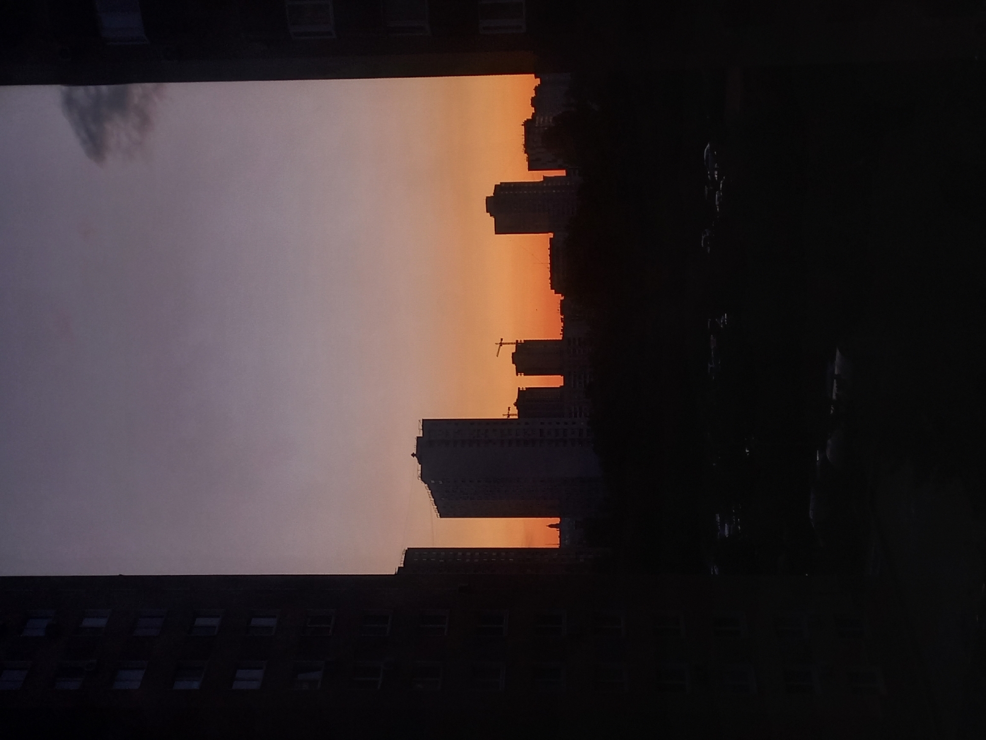 The image depicts a sunset over the city of Kyiv, Ukraine, viewed from a building window. The sky transitions from a pale purple-grey on the left to shades of orange and yellow towards the horizon. The silhouettes of several buildings are visible against the colorful sky, including some taller high-rises and a construction crane. In the foreground, parts of the building where the photo was taken are visible. The silhouette of other buildings, trees, and vehicles fill the right side of the frame.