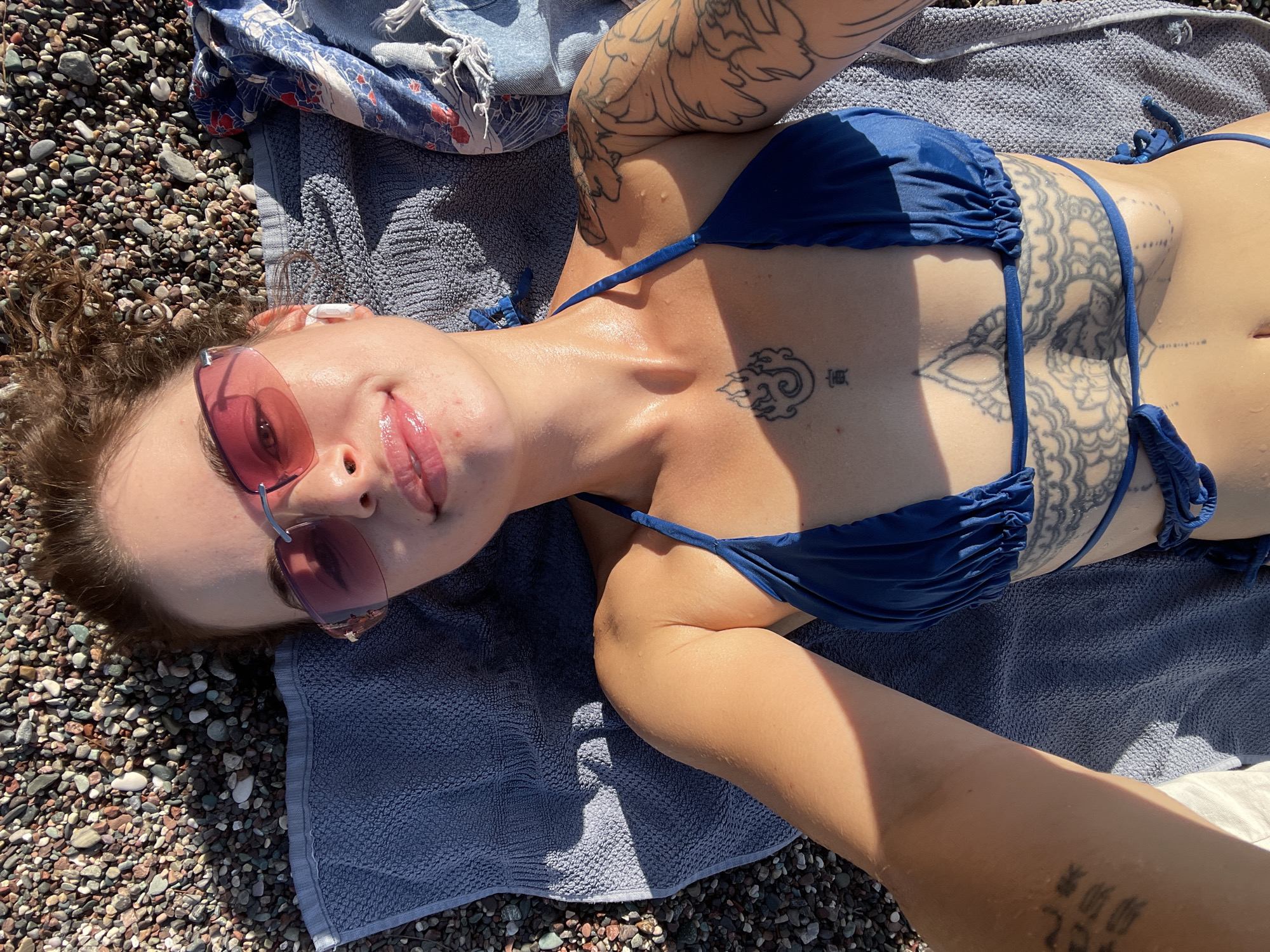 A woman lies on a beach in Rafailovići, Montenegro, on a towel placed on small pebbles. She is wearing a blue bikini and pink-tinted sunglasses. Her body is adorned with several tattoos, including intricate designs on her stomach and arms, and a flame-like symbol on her chest. She has white wireless earbuds in her ears. The sunlight casts a shadow across her body and the towel. A patterned cloth is draped to her right.