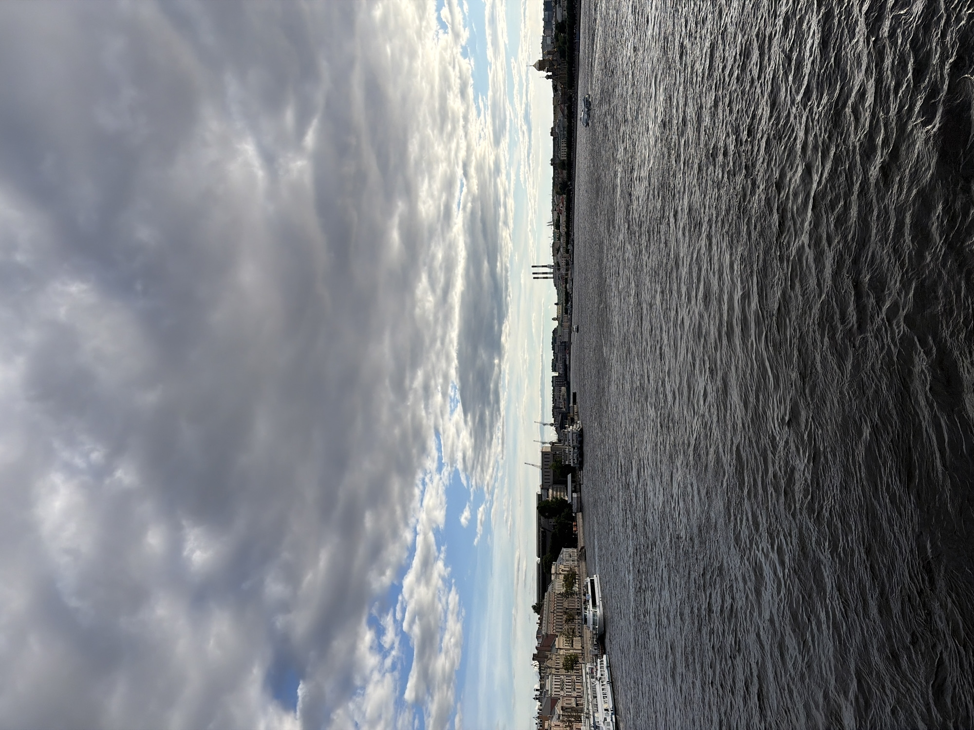 The image depicts a wide river with choppy, dark water in the foreground. Along the far bank, a cityscape stretches out under a dramatic sky filled with a mix of heavy grey clouds and patches of blue sky. Buildings with ornate facades and mansard roofs line the waterfront, suggesting a historical European city. Several white boats are docked along the embankment, and a few smaller vessels are visible on the water, including one creating a wake in the middle distance. Further down the river, industrial structures with tall smokestacks are visible, and in the far distance, a golden-domed building with a distinctive architectural style stands out against the horizon. The scene is captured from a slightly elevated viewpoint, likely from a bridge or another boat.