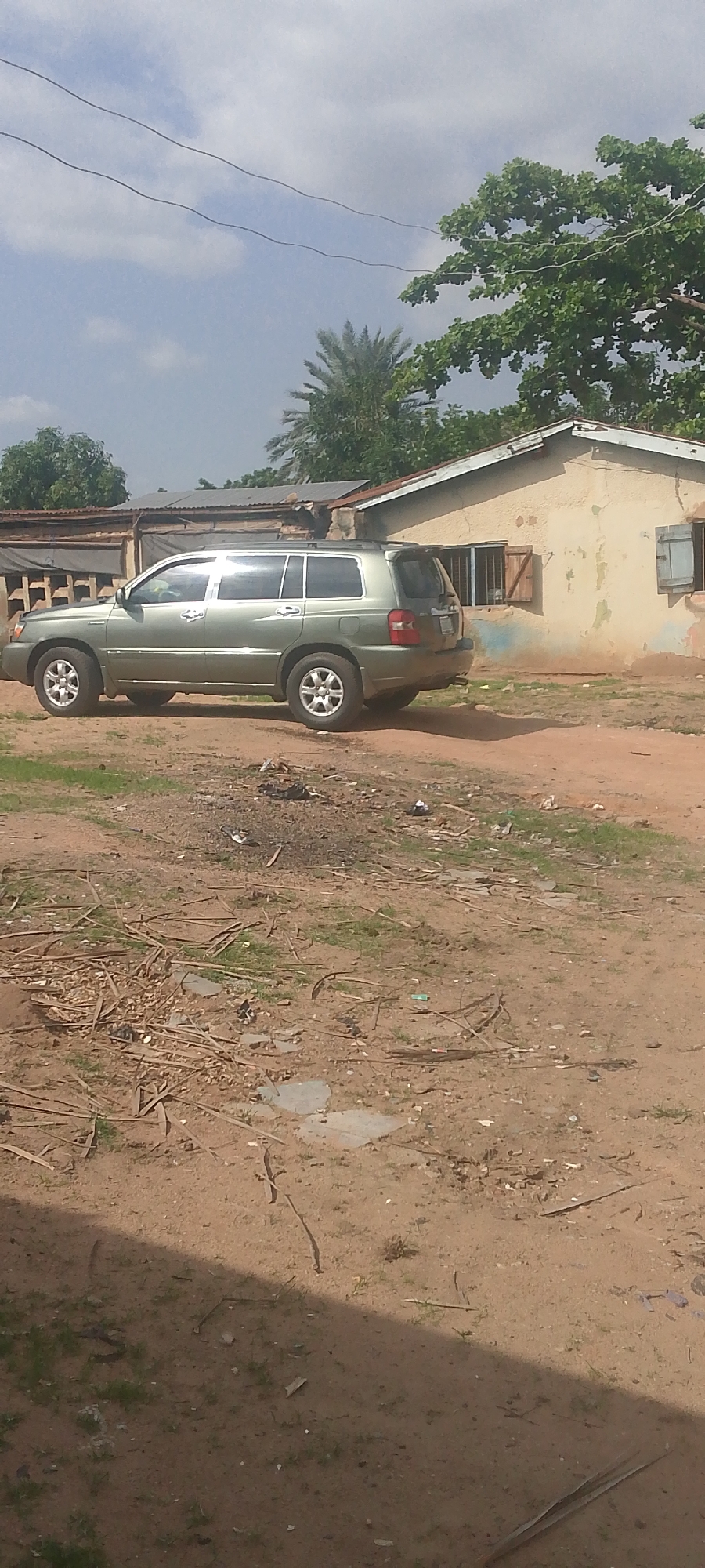 In Jalingo, Nigeria, a light green SUV is parked on a dirt road in front of a beige, stucco building with a corrugated metal roof. The building has wooden shutters on its windows, and the surrounding area is covered in dirt, dry leaves, and debris, including pieces of wood and what appears to be trash. Tall trees and a palm tree are visible in the background, and power lines crisscross the sky, which is partially obscured by clouds. The scene suggests an outdoor, rural or semi-urban setting.