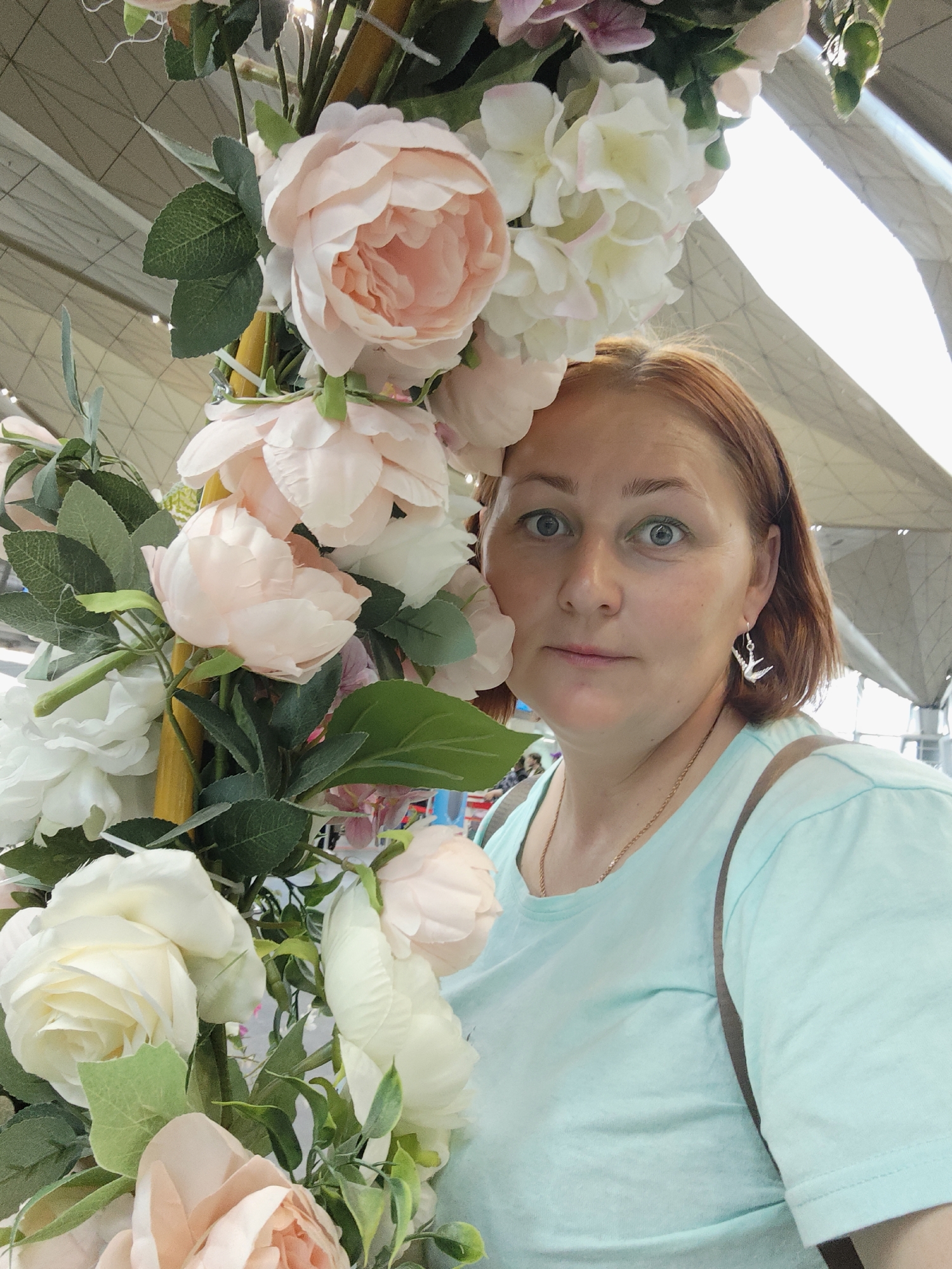 A fair-skinned woman with auburn hair, wearing a light blue t-shirt and a silver bird earring, is partially obscured by a large arrangement of light pink and white artificial flowers. Her eyes are wide and looking at the camera. She is positioned in the foreground, slightly to the right of center. The background appears to be an indoor space with a modern, curved ceiling structure, potentially an airport or exhibition hall, in Saint Petersburg, Russia. The lighting is bright, casting a soft glow on the scene.