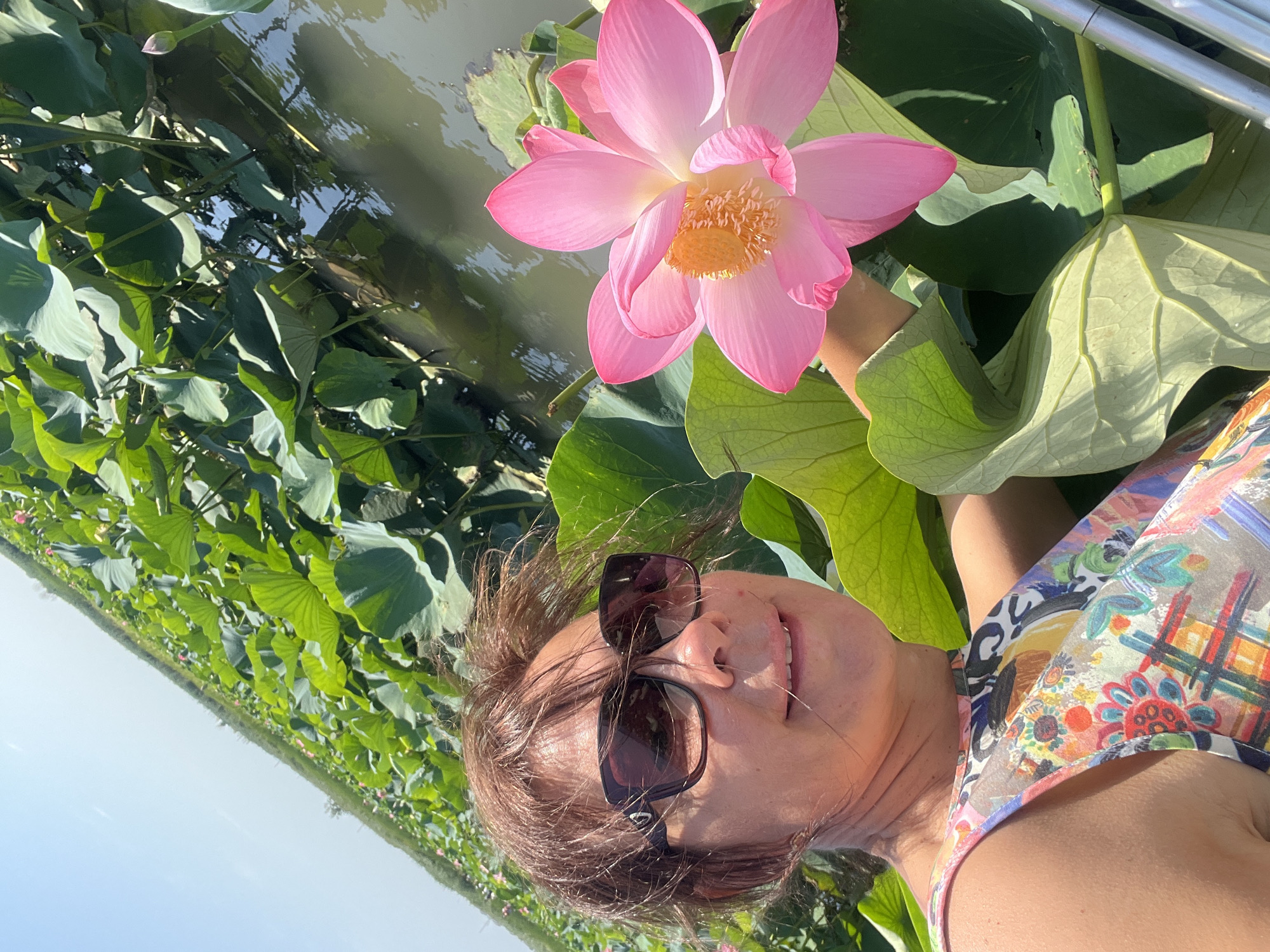 A woman with brown hair and sunglasses is in the foreground, smiling and looking slightly to the left. She is wearing a colorful, sleeveless dress. She is positioned in front of a body of water filled with large green lily pads and a large pink lotus flower. The lotus flower is in full bloom and its yellow center is visible. In the background, a vast expanse of lily pads stretches into the distance under a clear blue sky. The scene is consistent with a lotus farm, such as those found in Golubitskaya, Russia. The woman appears to be on a boat or platform, as indicated by the silver railing visible on the right side of the image.