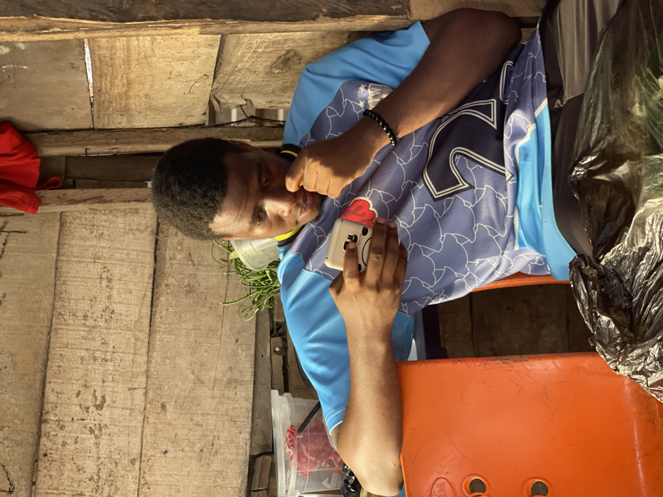 The image displays a young man in what appears to be a rustic or makeshift indoor setting, captured in a moment of quiet contemplation or focus. The original image is oriented sideways (rotated 90 degrees counter-clockwise), but the description will assume an upright perspective.
The central figure is a young man with dark skin and short, dark, curly hair. He is wearing a light blue short-sleeved t-shirt or sports jersey, which has a darker blue pattern (possibly stylized shapes or leaves) across the chest and shoulders, and a large white number "12" prominently displayed on the front. The collar is dark blue with a thin yellow stripe. On his left wrist, he wears a black beaded bracelet.
He is holding a light-colored smartphone in his right hand. The phone is encased in a distinctive white cover featuring a red ice cream cone design on its upper part and a simple, drawn-on cartoon face (two dots for eyes, a curved line for a mouth) on its body. His gaze is directed downwards and slightly to his right, suggesting he is looking at something off-frame or intently at his phone. His left hand is raised, with fingers slightly curled, resting near his chin or mouth, conveying a pensive or relaxed posture.
The background consists primarily of rough, untreated wooden planks, forming what appears to be a wall or part of a simple structure. The wood is light-colored and shows visible grain and texture, with some gaps between the planks.
Several other objects are visible in the scene:
* An orange plastic stool or chair occupies the lower right foreground, with the man's right forearm resting on it.
* To the right of the man, a crumpled black plastic bag is visible.
* Behind him, to his left, is a translucent white plastic container holding some green leafy vegetation, possibly herbs or vegetables.
* Further to the left, a red fabric item, possibly a piece of clothing, is partially visible against the wooden wall.
* Another clear plastic container is visible on the ground or a lower surface beneath the green vegetation.
The lighting suggests it is daytime, with natural light entering from the right side of the frame (when upright), casting subtle shadows and highlighting parts of the man's face and arms. The overall atmosphere is unpretentious and casual, focusing on the man's absorbed state. No specific text is clearly legible other than the number "12" on his jersey and the simple face on the phone case.