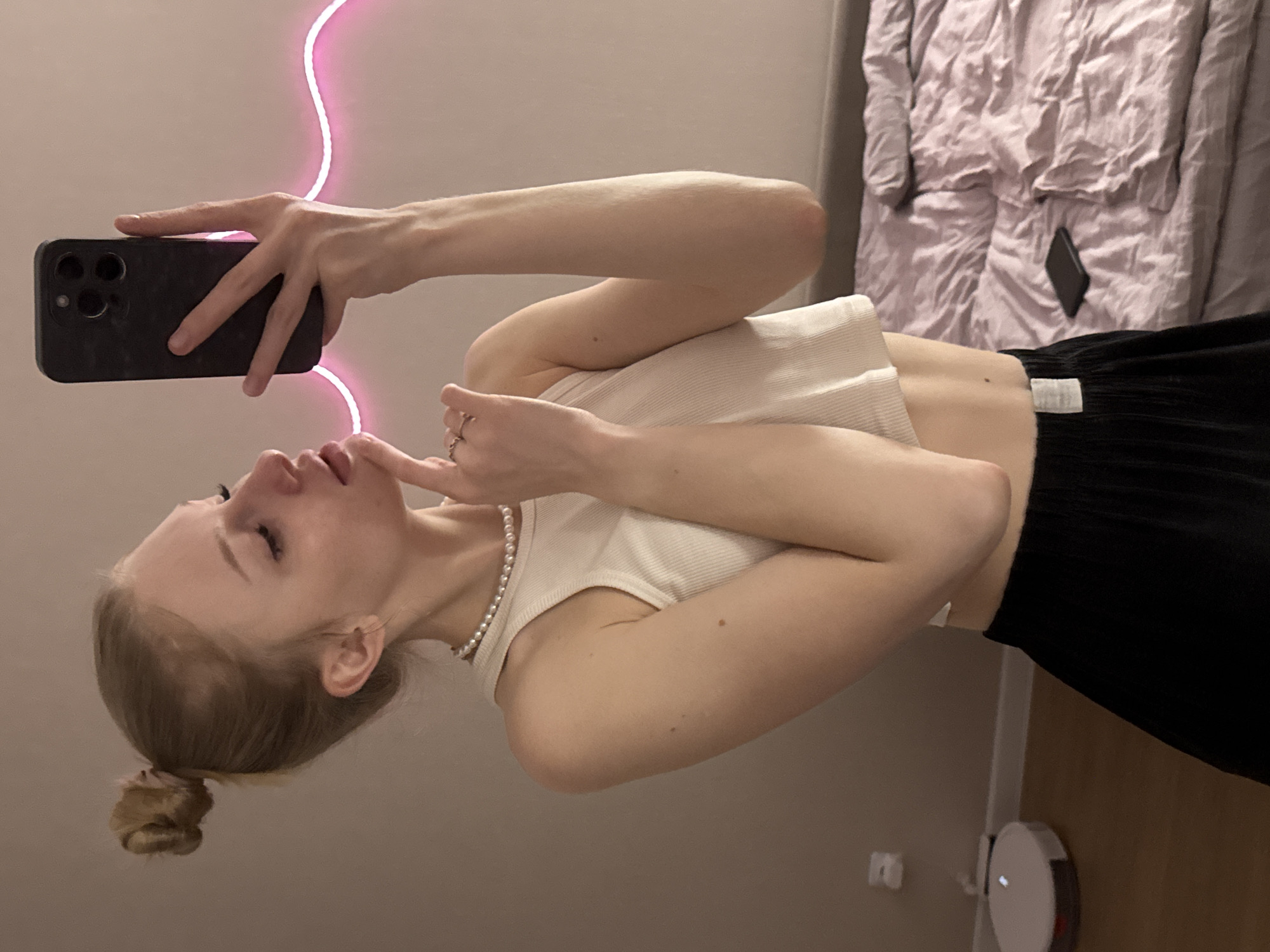 A young female with light skin and blonde hair tied in a high bun is depicted taking a mirror selfie in an indoor residential setting, identified as the city of Perm, Russia. She is holding a dark-colored smartphone, likely an iPhone, in her right hand, which partially obscures the right side of her face. Her head is tilted slightly back and to her right, with her eyes partially closed. Her left hand is raised, with the index finger touching her chin and a ring visible on her fourth finger.
She is wearing a white ribbed crop top, exposing her midriff, and a single-strand pearl necklace. Black wide-leg pants with a light-colored tag on the left waistband cover her lower body. A small, dark mole is visible on her upper left arm/shoulder, and another on her lower left torso.
The background features a light-colored, possibly beige or pale pink, wall. In the upper right background, a wavy pink LED strip light is affixed to the wall. To the right, a bed with wrinkled light purple bedding and pillows is visible, with a second dark smartphone placed face down on the bed. In the lower-left corner of the image, a white circular robot vacuum cleaner is positioned on a wooden floor, near a white baseboard.