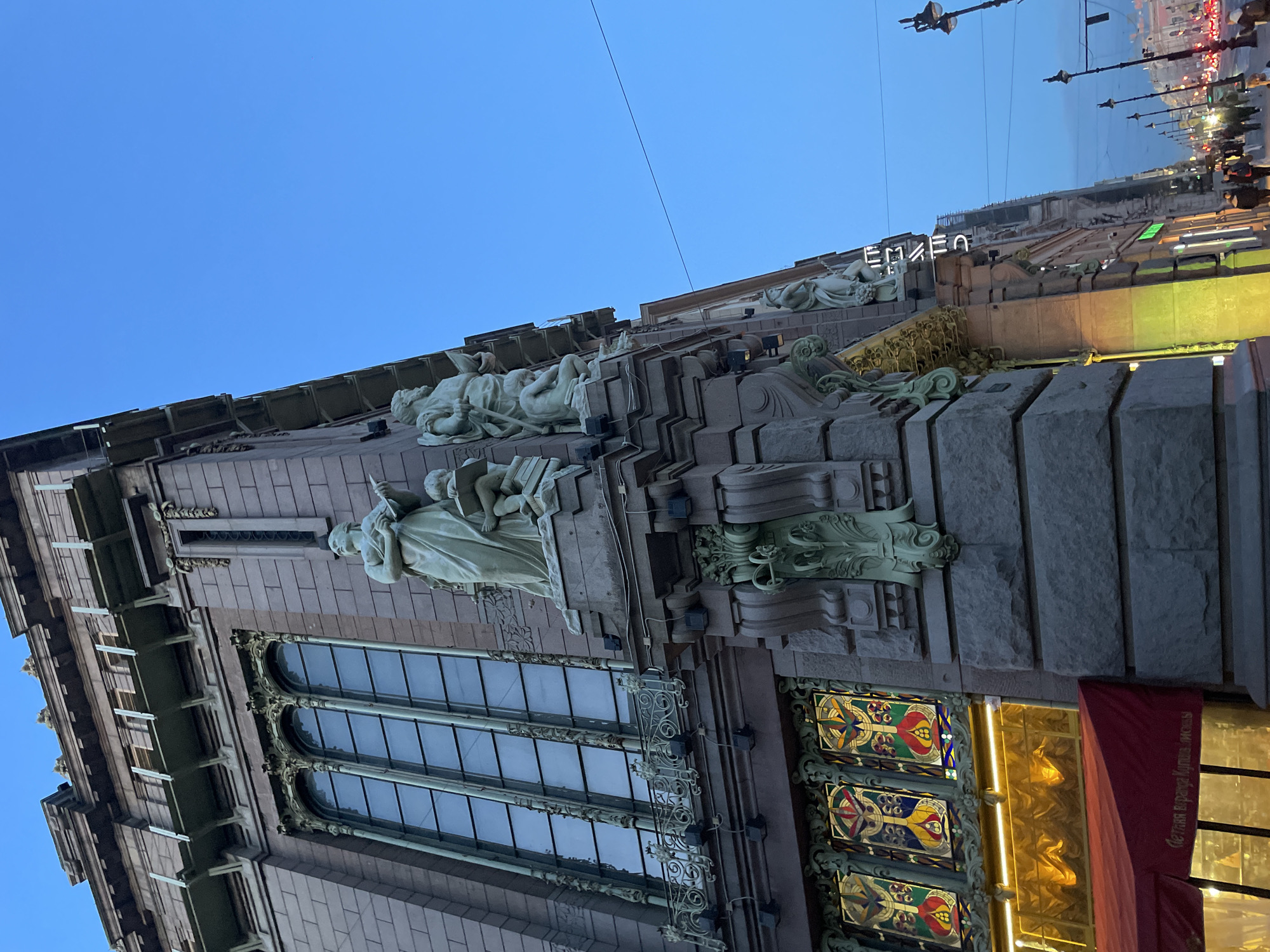 An ornate building facade, viewed from a low angle, dominates the frame against a clear, deep blue sky, indicating dusk or early evening. The multi-storied structure is rich in detail, featuring numerous classical-style sculptures of human figures. Some figures are reclining on ledges, while others stand upright, one appearing to hold a book. These allegorical sculptures are integrated into various levels and decorative projections of the building.
Below the main set of sculptures, large arched windows with intricate metalwork can be seen. Further down, at street level, there are decorative panels resembling stained glass, featuring colorful, geometric patterns in red, yellow, and blue. Next to these, a golden-lit entrance area is visible, partially obscured by a red banner with partially visible Cyrillic text.
At the upper part of the building, a prominent neon sign spells out "ЕЛИСЕЕВ" (ELISEEV) in bright white letters, illuminating the structure. In the background, across the street, blurred outlines of other buildings, numerous streetlights, and distant vehicle lights suggest a busy urban setting. The overall impression is one of grand, historical Art Nouveau architecture characteristic of Saint Petersburg.