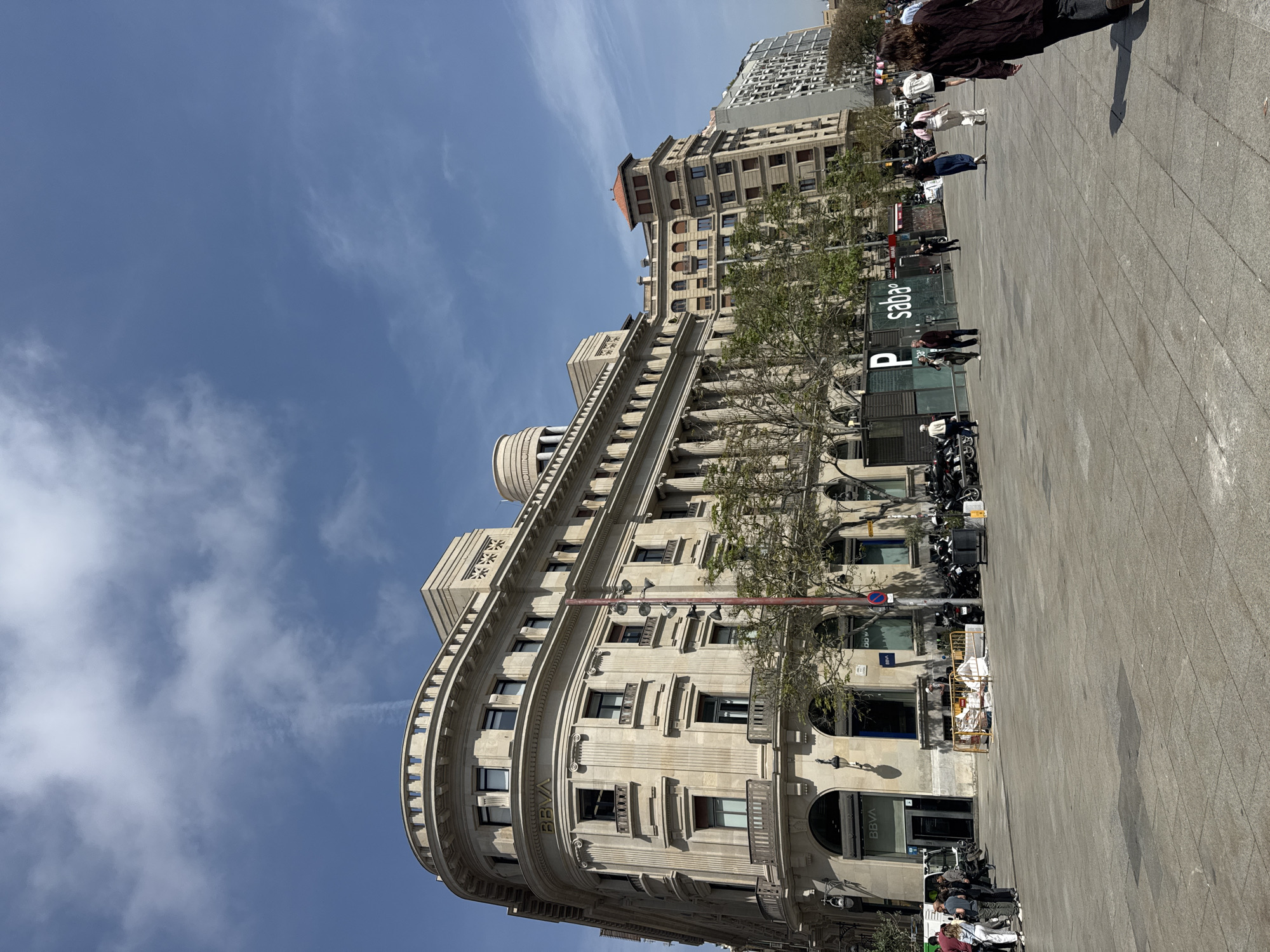 This media file captures a bright, sunny daytime scene of a bustling urban square in Barcelona, Spain. The prominent subject is a grand, multi-story building with a curved, ornate stone facade, featuring classical architectural elements such as pilasters, cornices, balustraded balconies, and arched windows. The building houses a BBVA bank, with the "BBVA" logo visible multiple times on its lower floors.
To the right and in the foreground, a wide, paved plaza or pedestrian area stretches out. Several individuals are scattered across this space, some walking, others standing. A notable feature is a "Saba P" sign, indicating a parking entrance, near which several motorcycles and scooters are parked. Yellow temporary barriers are also visible at the base of the ornate building. In the background, beyond the main building, another taller, more modern structure can be seen. The sky is clear and blue with scattered wispy white clouds, suggesting fine weather. The overall impression is one of typical urban activity on a pleasant day.