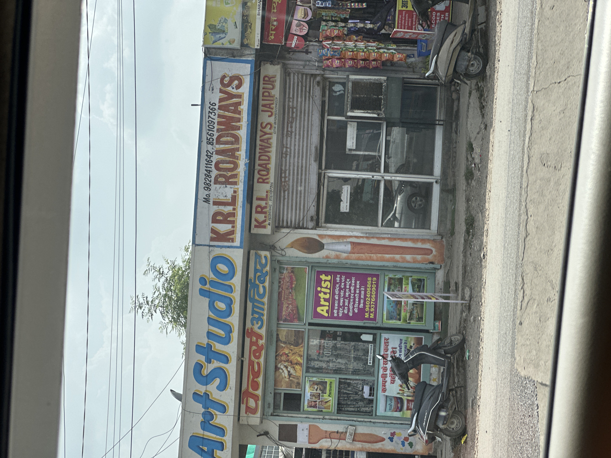The image captures a street-level view of a commercial establishment in Jaipur, India. The facade of the building features multiple shopfronts. Prominently displayed is a sign for "Art Studio" in large blue letters with a yellow outline. Adjacent to it, another sign reads "K.R.L. ROADWAYS" in red lettering. Below these, a shop sign in Hindi states "Posters Artist". The shop window displays various artworks and portraits, including what appear to be traditional Indian paintings. A large graphic of a paintbrush is visible on the right side of the shop. Another sign on the shop states "Artist" and lists services related to painting and portraits.
Further to the right, a shop with a rolling shutter displays merchandise, possibly snacks and small packaged goods, hanging from racks. A sign above this shop reads "K.R.L. ROADWAYS JAIPUR".
In front of the shops, a dark-colored scooter is parked on the side of the road. Another lighter-colored scooter is parked further down the street. The road surface appears to be asphalt, with some cracks and debris. The environment suggests a typical urban setting in India, with overhead power lines visible against a partly cloudy sky. The overall scene depicts a commercial area with a focus on art and transportation services.
