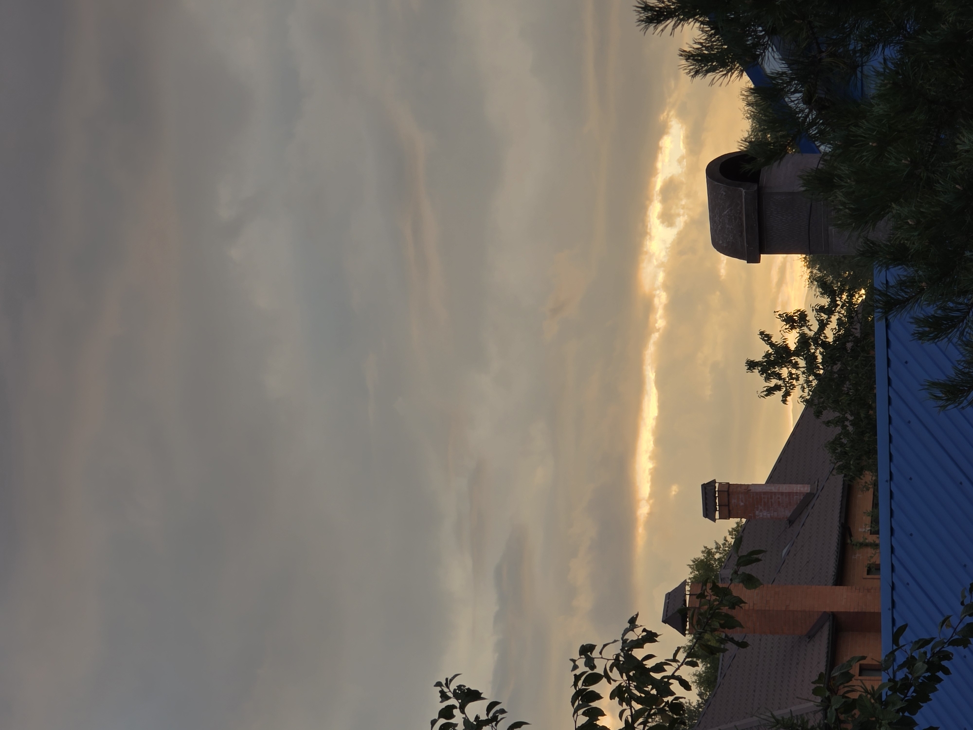 The image depicts a residential area with houses and rooftops under a cloudy sky at what appears to be dusk. The sky is filled with diffused light, with streaks of warm orange and yellow suggesting the last rays of sunlight. Several chimneys are visible on the roofs, indicating an active heating system in the homes. Trees and foliage frame the scene, adding a natural element to the urban landscape. The presence of these elements suggests a quiet evening in a suburban or rural setting. The mention of Речицы, Russia, places the image in a specific geographical context within the Russian Federation.