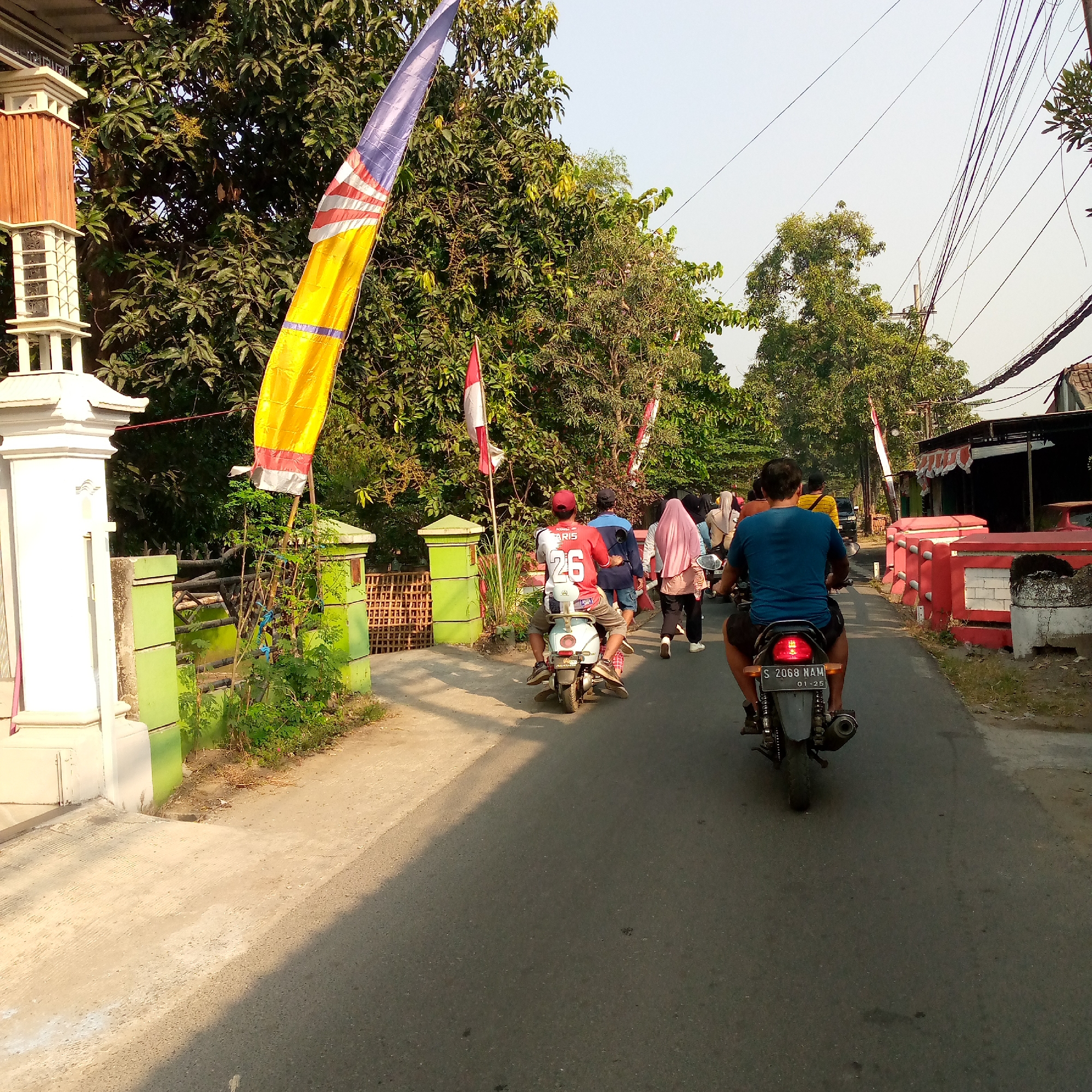 The image depicts a street scene in Tambakagung, Indonesia, with several people on motorcycles and walking. In the foreground, two individuals are riding motorcycles, one in a blue shirt and black shorts, and another on a scooter wearing a red shirt with the number "26" on the back and a red cap. To the right of the road, there are red and white structures along the roadside, possibly part of a cemetery or a decorative boundary. A large yellow and blue flag is prominently displayed on the left side of the road, near a white pillar and a green fence. Trees line the street, and utility wires are visible overhead. Several other people are walking and riding motorcycles further down the road, suggesting a procession or a busy public area. The road appears to be asphalt, and the time of day seems to be late afternoon, given the shadows cast by the trees and the quality of light. The overall impression is that of a local community event or a typical day in a suburban Indonesian setting.