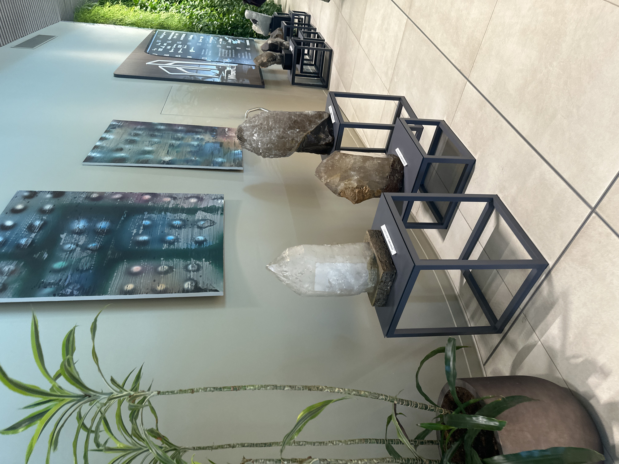 The image shows an indoor display of minerals and artwork. In the foreground, a spider plant in a dark pot is partially visible. Behind the plant, on a light-colored wall, are two rectangular artworks with abstract, colorful patterns, resembling liquid or nebulae. To the right of the artworks, a row of modern, dark metal cube-shaped stands display large mineral specimens. The minerals appear to be quartz, with one large, clear crystal prominently featured, wrapped in plastic. Other specimens are translucent and brownish, some with visible facets. The background consists of a tiled floor and a wall, suggesting a museum, gallery, or a modern exhibition space. The lighting is bright and even, indicating an indoor setting, possibly during the daytime. A small piece of text is visible on a label attached to one of the mineral displays, but it is too blurry to read. The overall impression is one of a clean, minimalist exhibition focused on natural geological formations and abstract art. The setting is likely a public space in Sofia, Bulgaria.