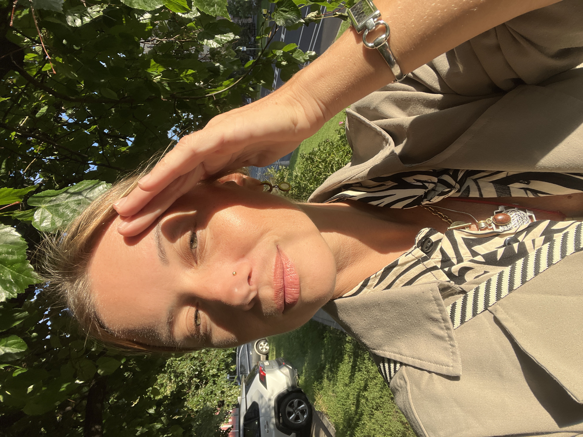 A woman is taking a selfie outdoors in Moscow, Russia. She is wearing a beige trench coat over a patterned shirt and has her hand on her forehead, as if shielding her eyes from the sun. She has a nose piercing and is wearing jewelry on her wrist and around her neck. In the background, green trees and a street with cars are visible. The sun is shining, indicating it is daytime. The image appears to be taken on a clear, sunny day. The woman's expression is relaxed and she seems to be enjoying the weather. There is no visible text in the image.