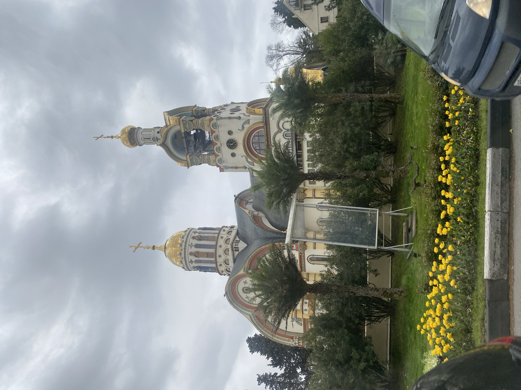 The image captures a church situated in Sochi, Russia, under an overcast sky. The architectural style is notably Russian Orthodox, featuring golden domes and a bell tower. The church's facade is adorned with decorative elements, including arches and patterns. Lush greenery, including palm trees and flowering plants, surrounds the church, creating a park-like setting. A vehicle is partially visible on the side of the image, suggesting the location is adjacent to a street.