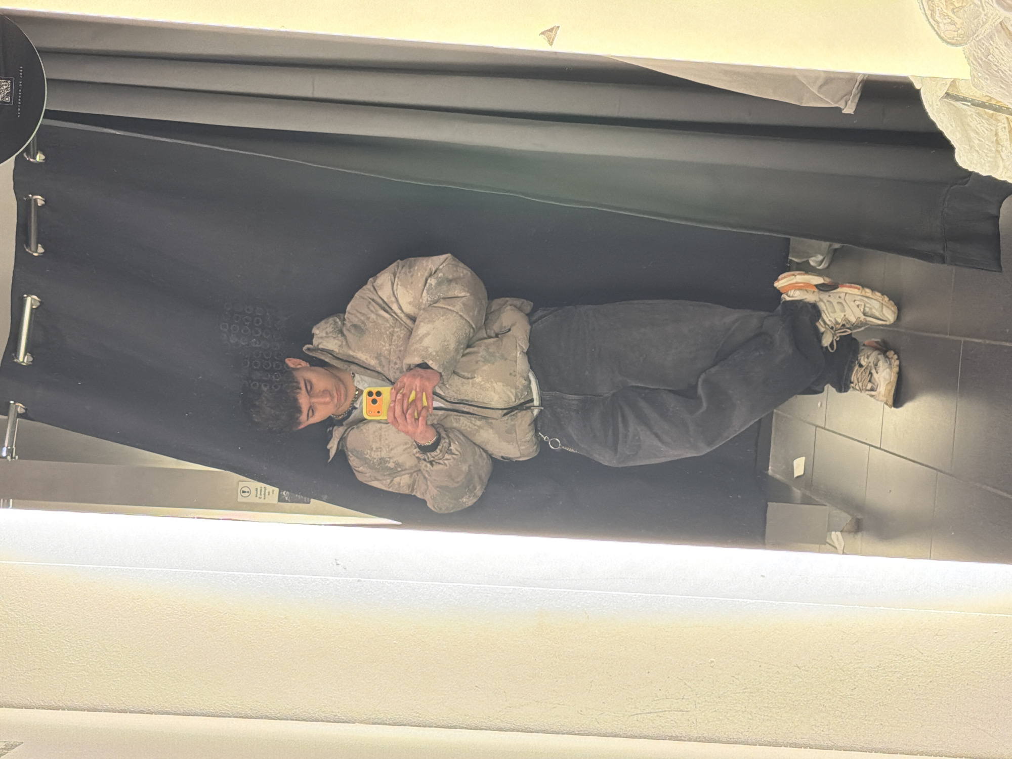 A young man is lying down in a changing room. He is holding a yellow smartphone. The changing room has a black curtain. The location is likely in a retail establishment, but the city (Sofia, Bulgaria) cannot be ascertained from the image alone.